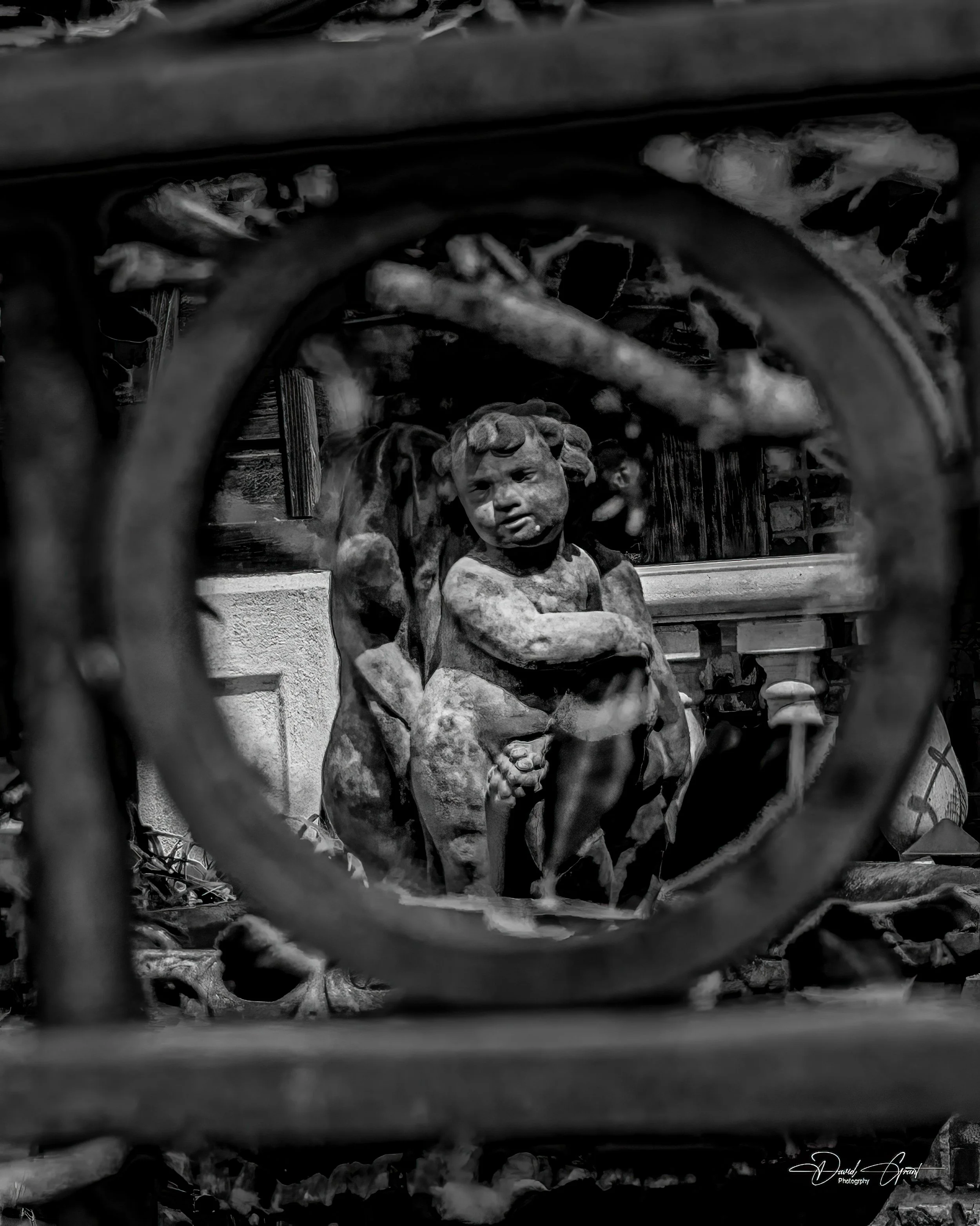 Black and white photo of a small statue of a child with animal-like features, sitting with arms crossed. The statue is viewed through a circular opening in a decorative iron fence. Background includes a wooden fence and foliage.