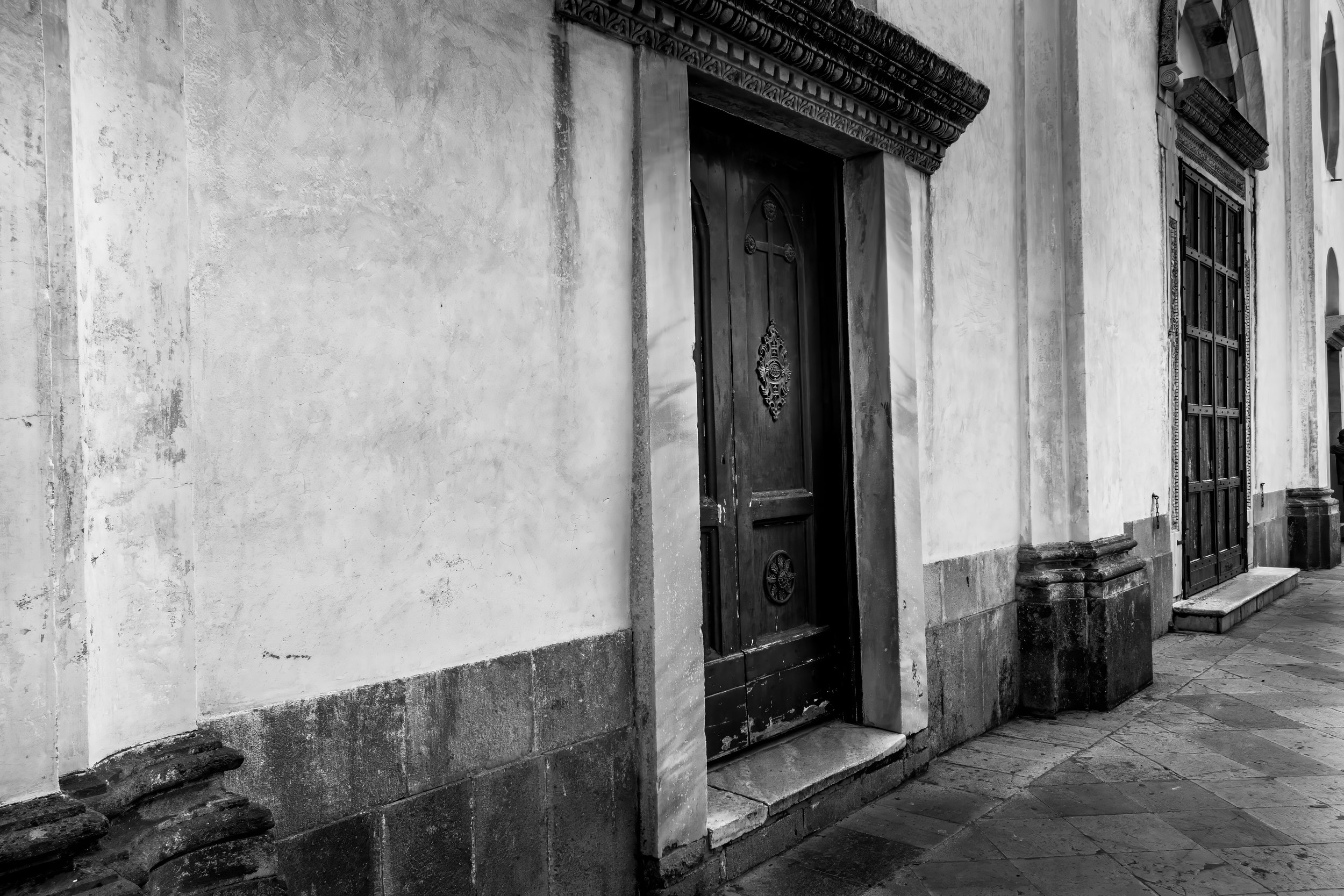 Black and white photo of an old building with a wooden door and a large window with multiple panes, both framed with ornate molding. The building's wall has a weathered appearance.