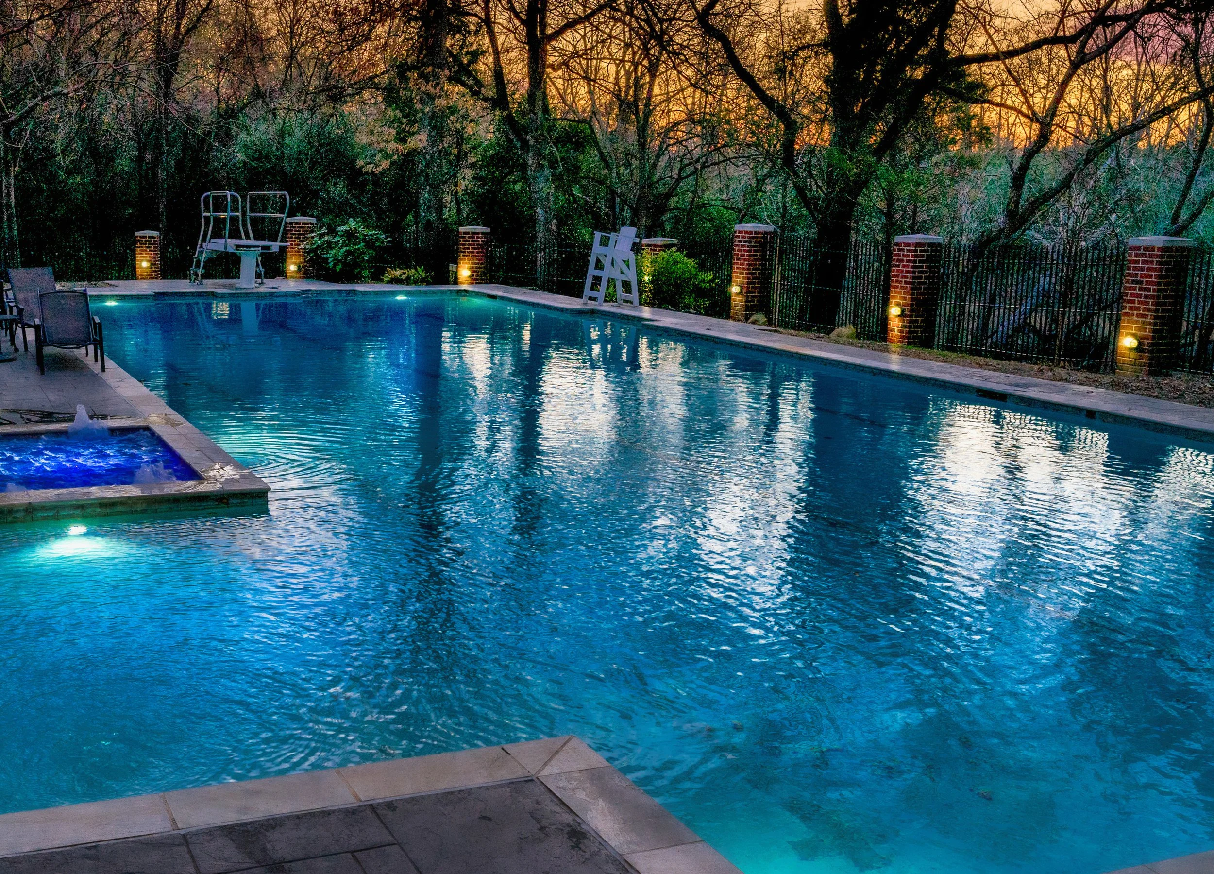 A backyard swimming pool with a smaller hot tub in the corner, surrounded by a patio with chairs and a sunset sky, trees, and a fence.