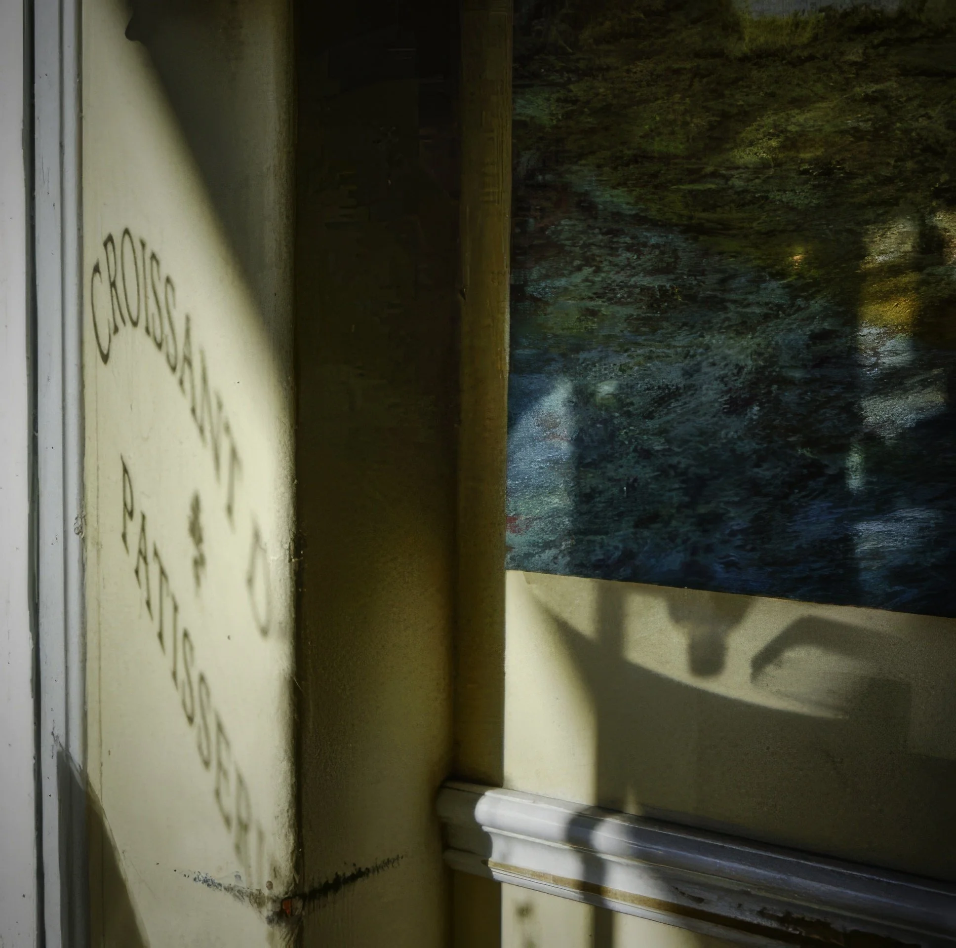 Partial view of a yellow wall with the words 'CROSSGATE PAINTING' slightly visible, with a shadow cast on the wall, a wooden frame, and a blue abstract painting hanging on the wall.