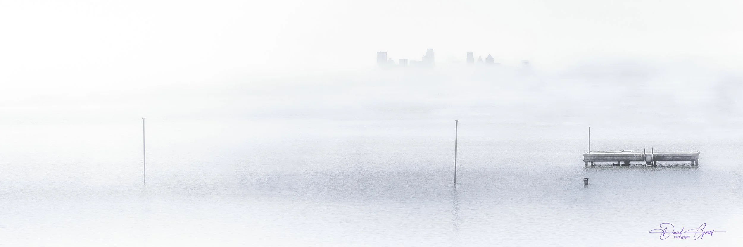 A foggy waterfront scene with a dock extending into calm water, positioned to the right. Tall poles are in the water, and a city skyline is faintly visible in the background through the fog.