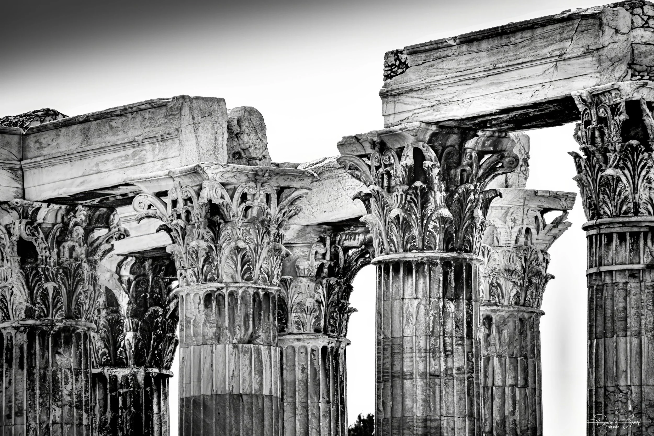 Ancient stone ruins of Greek or Roman columns with ornate Corinthian capitals, some broken and weathered, in black and white.