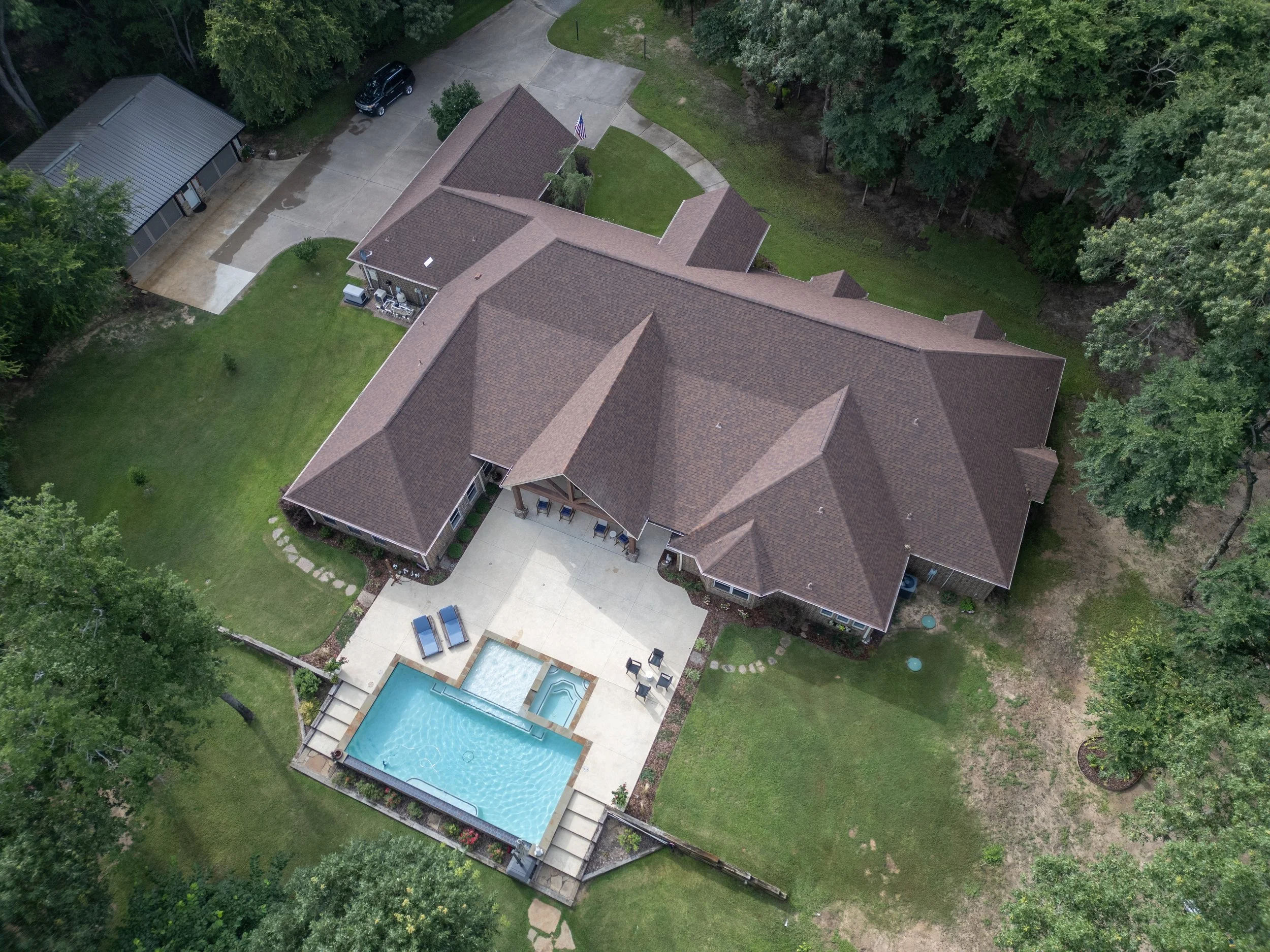 Aerial view of a large house with a brown roof, a backyard with a swimming pool, patio furniture, and surrounded by trees and greenery.