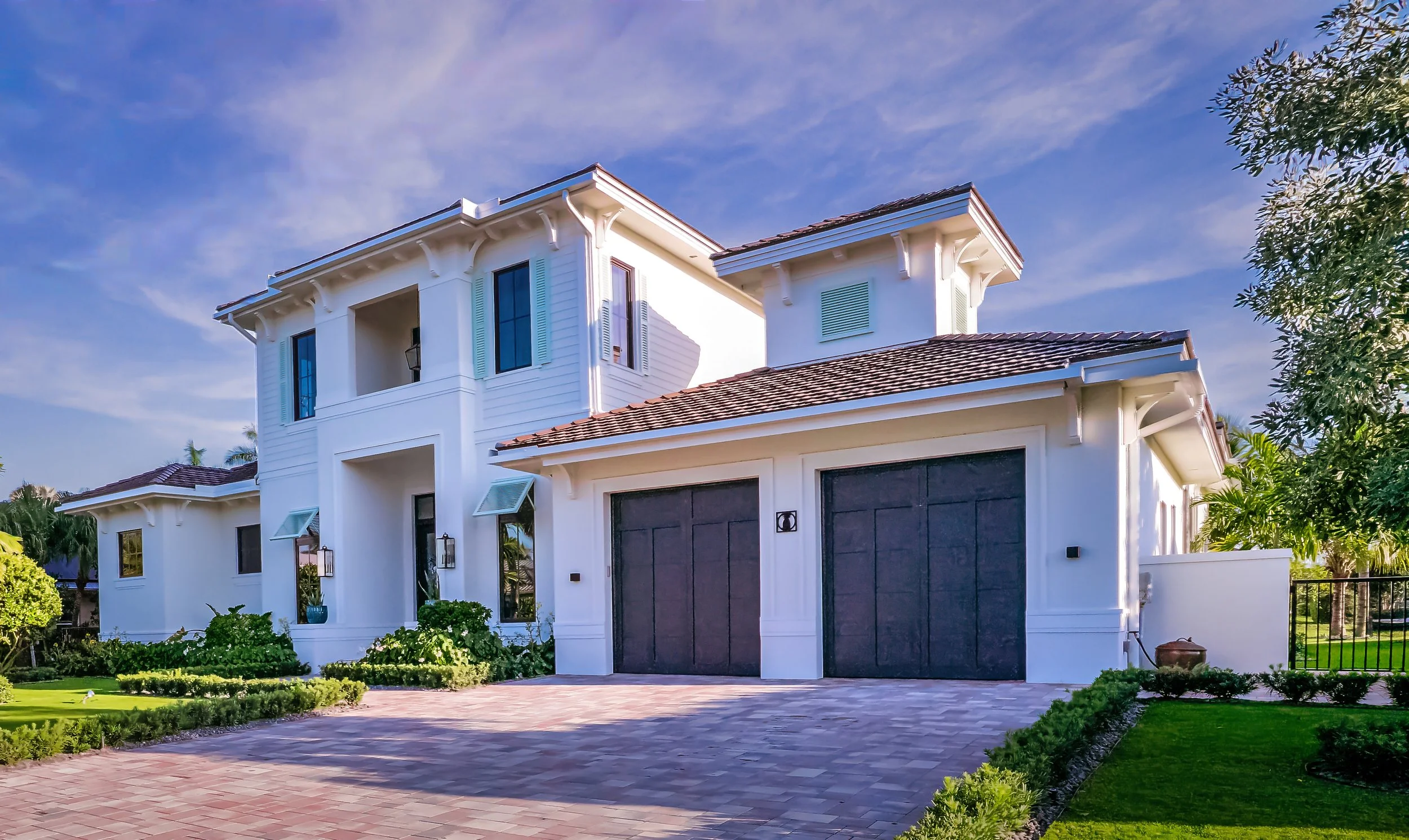 Front view of a modern white house with a brown tiled roof, black garage doors, and a paved driveway, surrounded by lush green landscaping and trees under a blue sky.