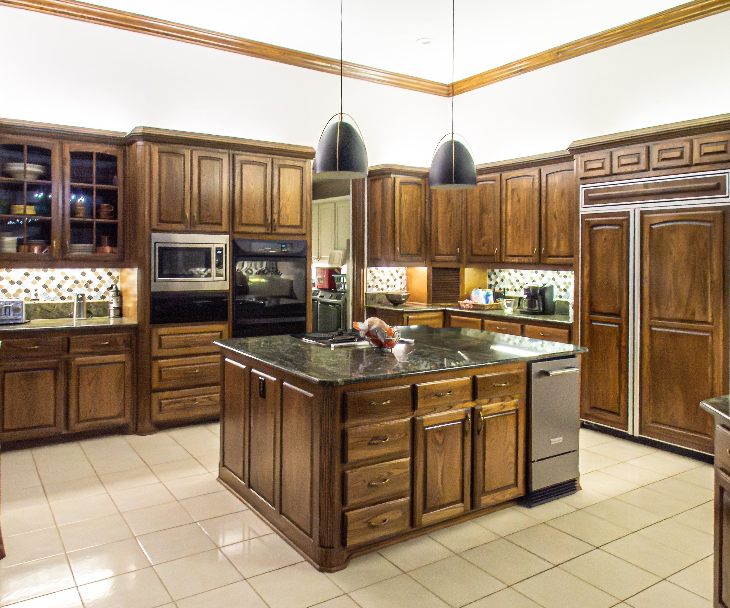 Wooden kitchen with black granite countertops, island, microwave, oven, refrigerator, and tiled floor.
