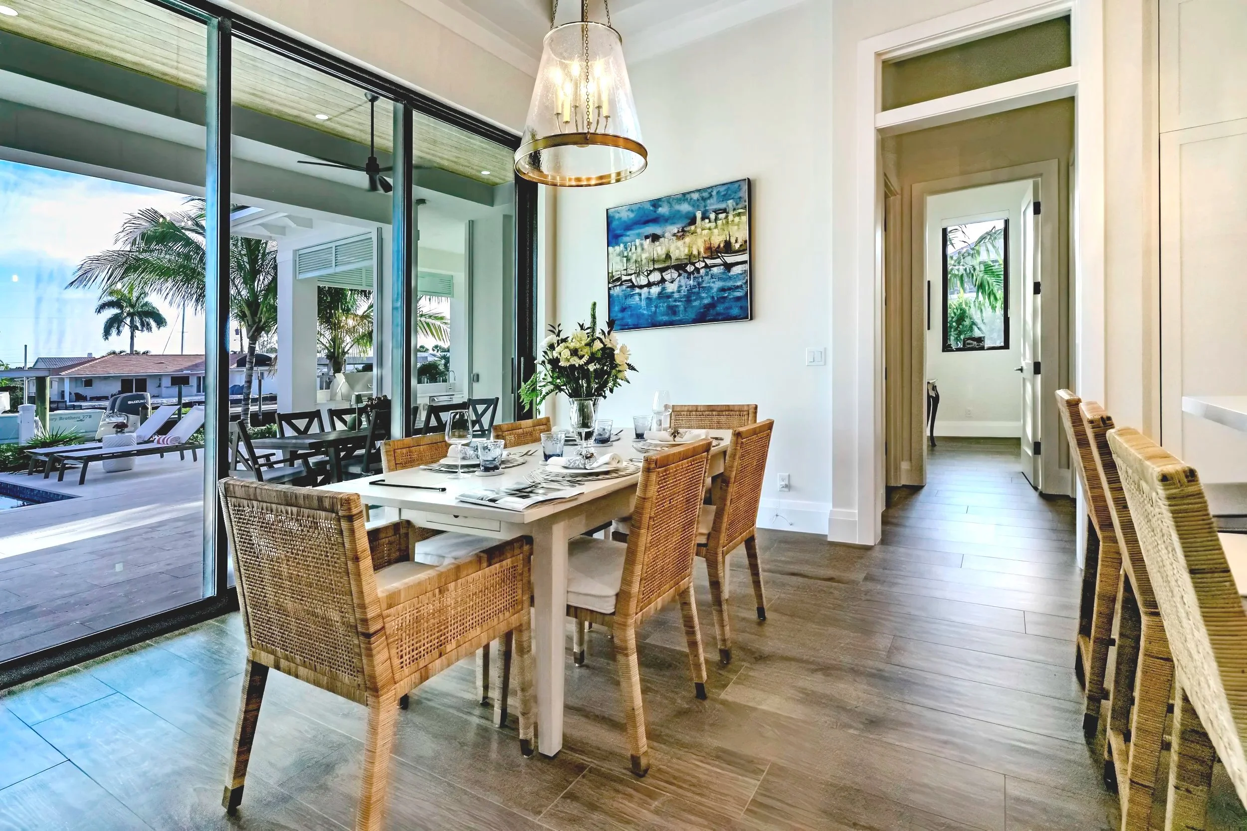 Dining area with a table set with glassware and a floral centerpiece, open to an outdoor patio with lounge chairs and palm trees visible through large glass sliding doors.