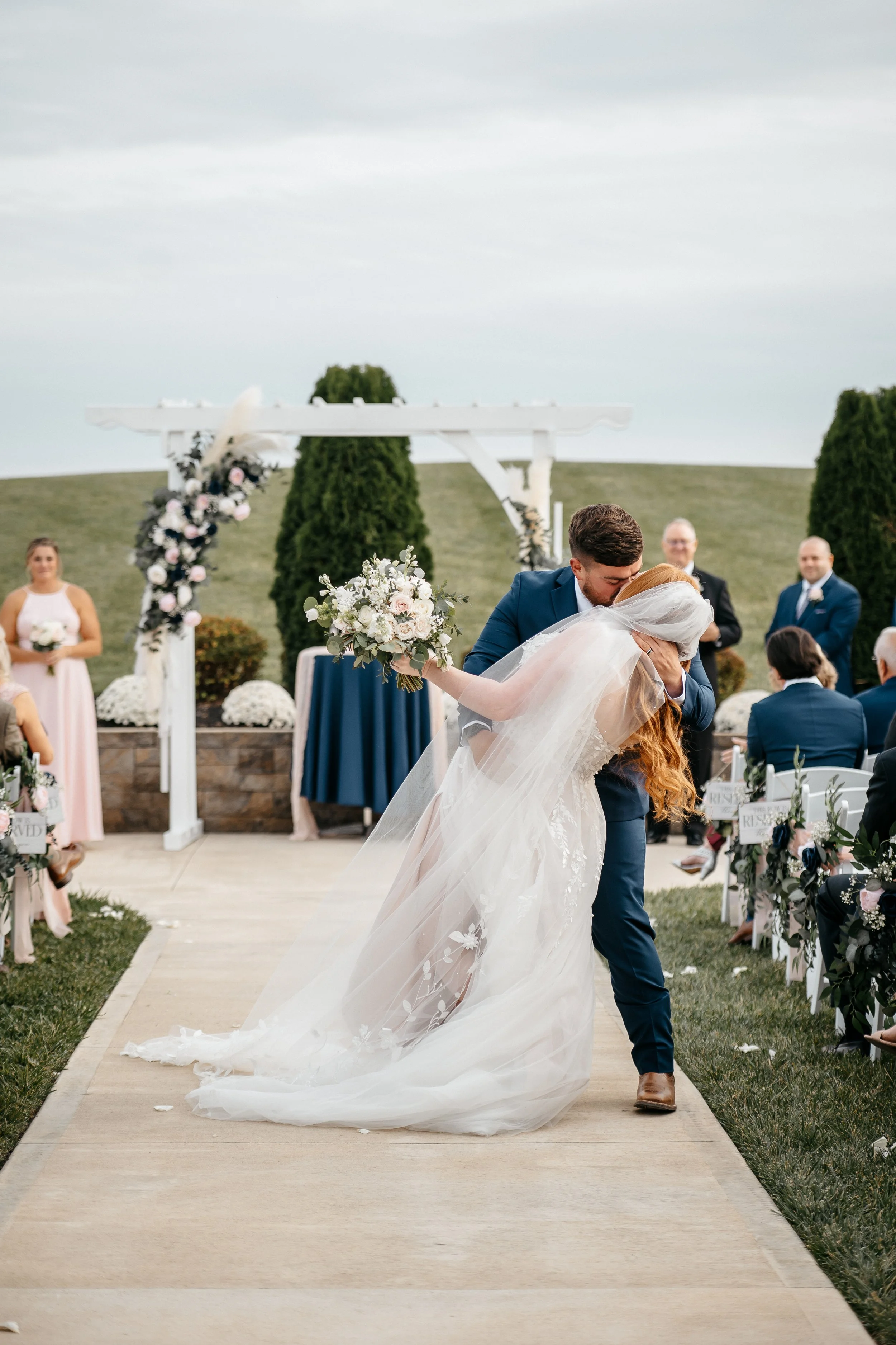 A Dayton Ohio bride and groom share a kiss in the isle at their outdoor wedding ceremony, with guests seated on either side and a decorated arch in the background.