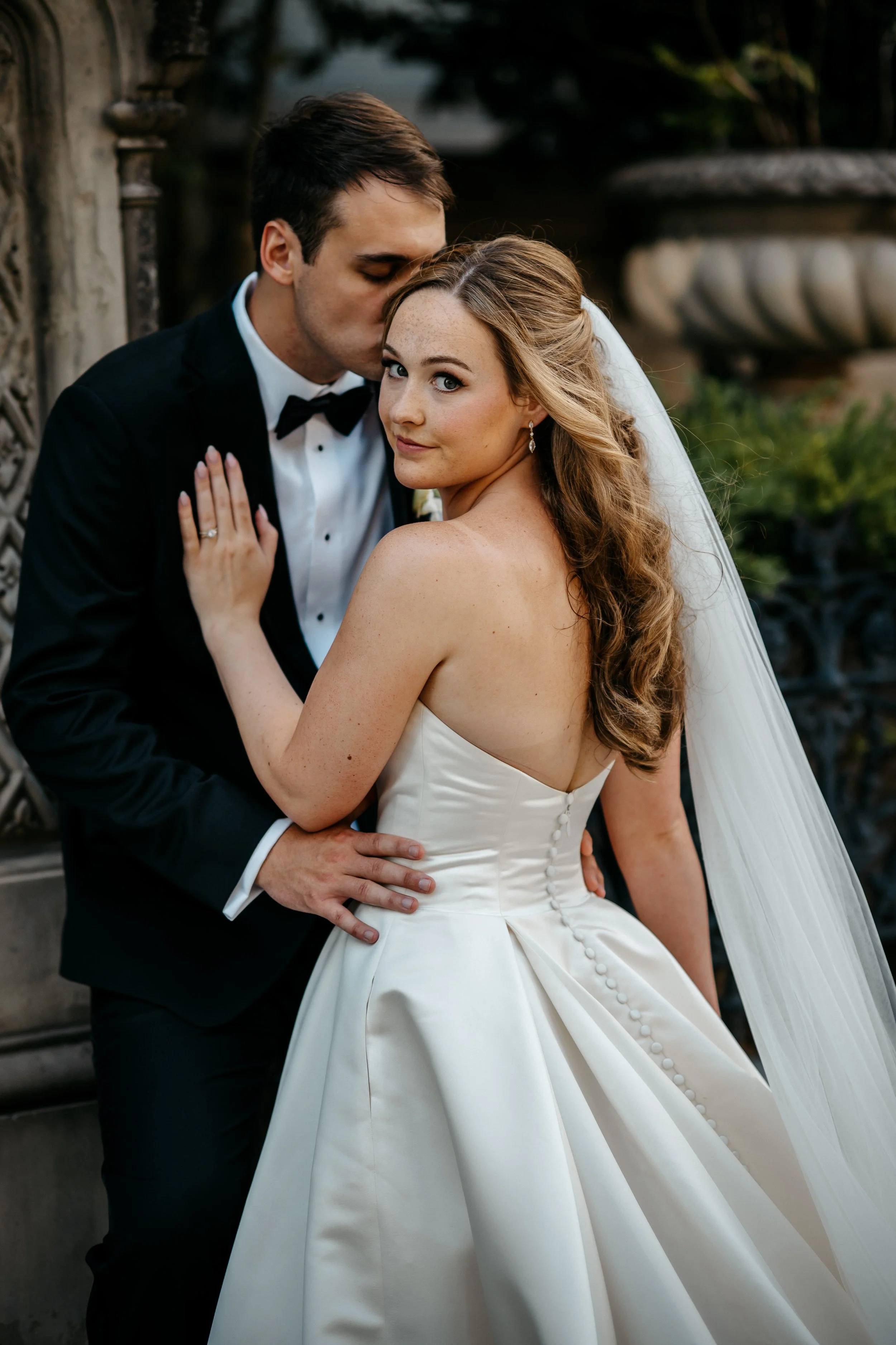 A Cincinnati Ohio bride and groom in wedding attire posing together outdoors. The groom is kissing the bride's forehead, and she is looking at the camera with a gentle smile.