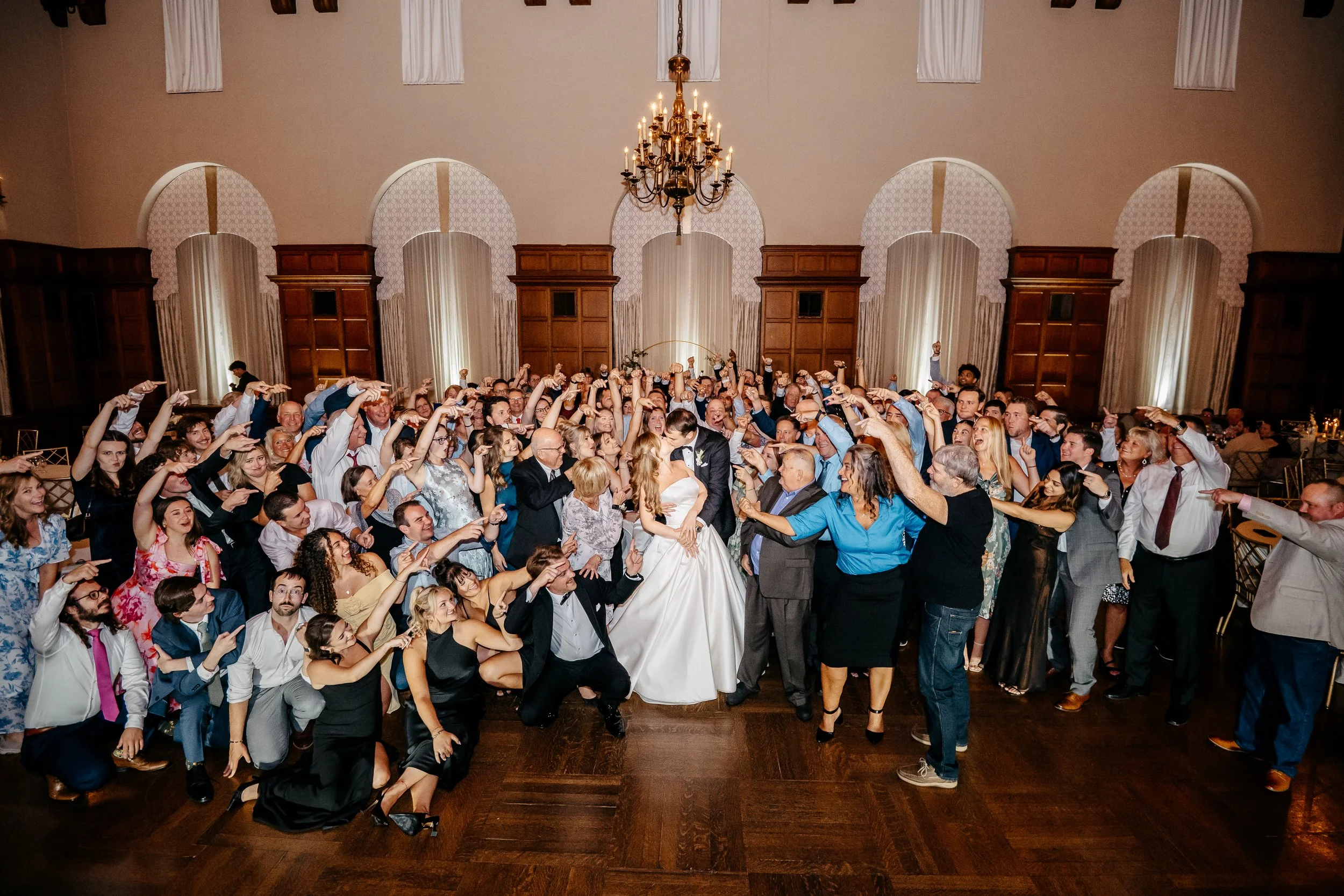 A large Cincinnati Ohio wedding reception with the bride and groom kissing, surrounded by many guests dancing and celebrating in a grand hall with chandelier and high ceilings.
