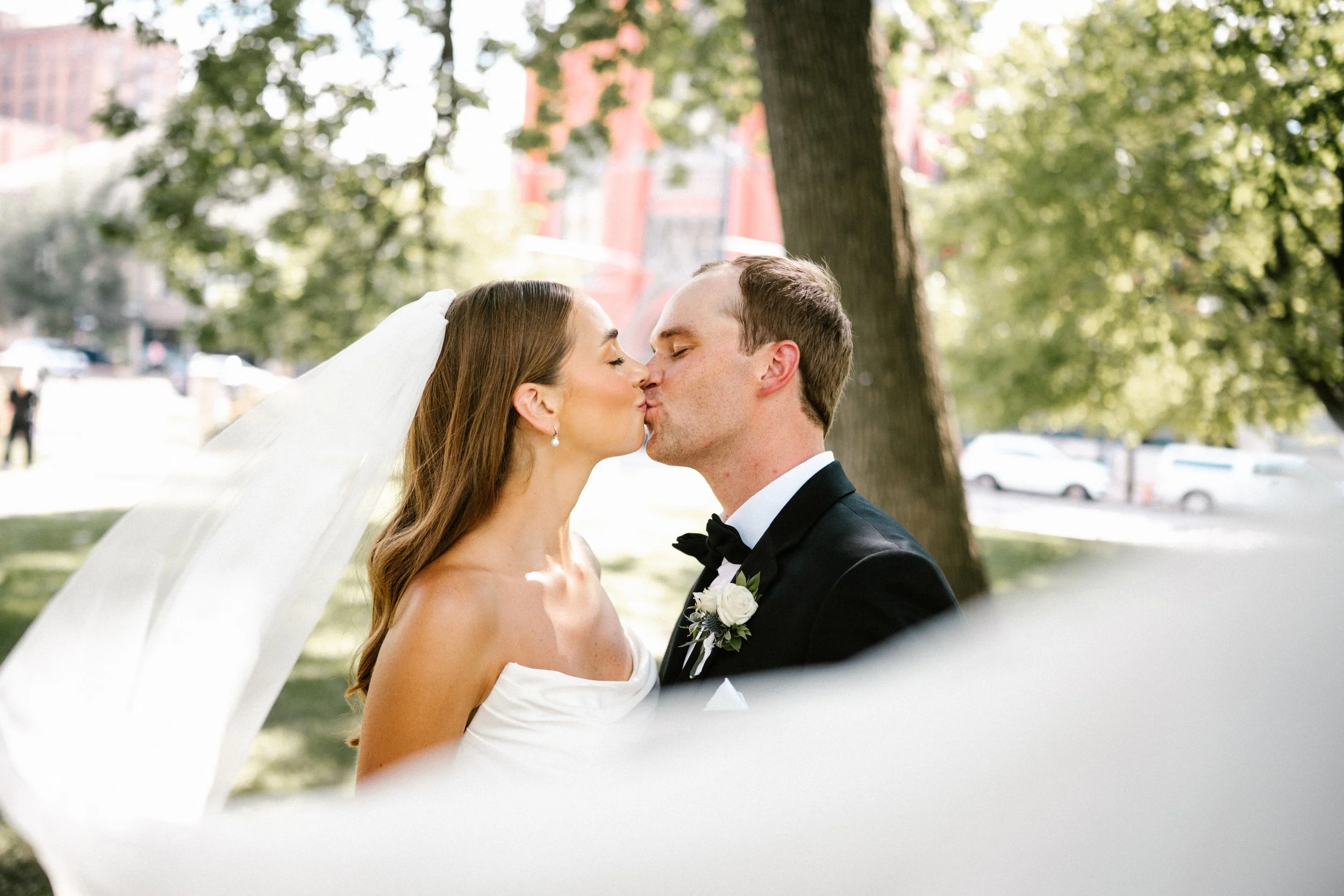 A Cincinnati Ohio bride and groom sharing a kiss outdoors with vail blowing in the wind during their wedding, with trees and a city building in the background.