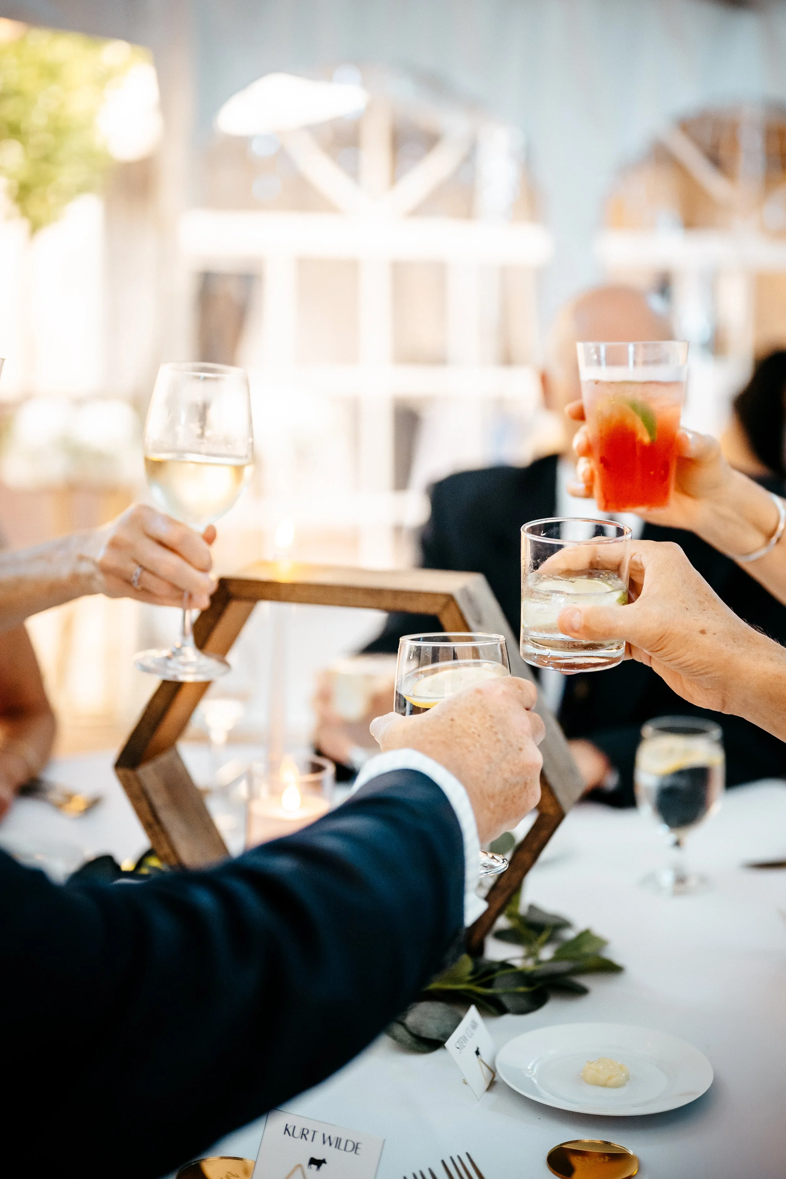 People clinking glasses with wine and cocktails at a table during a wedding celebration, with a decorative centerpiece and candles.