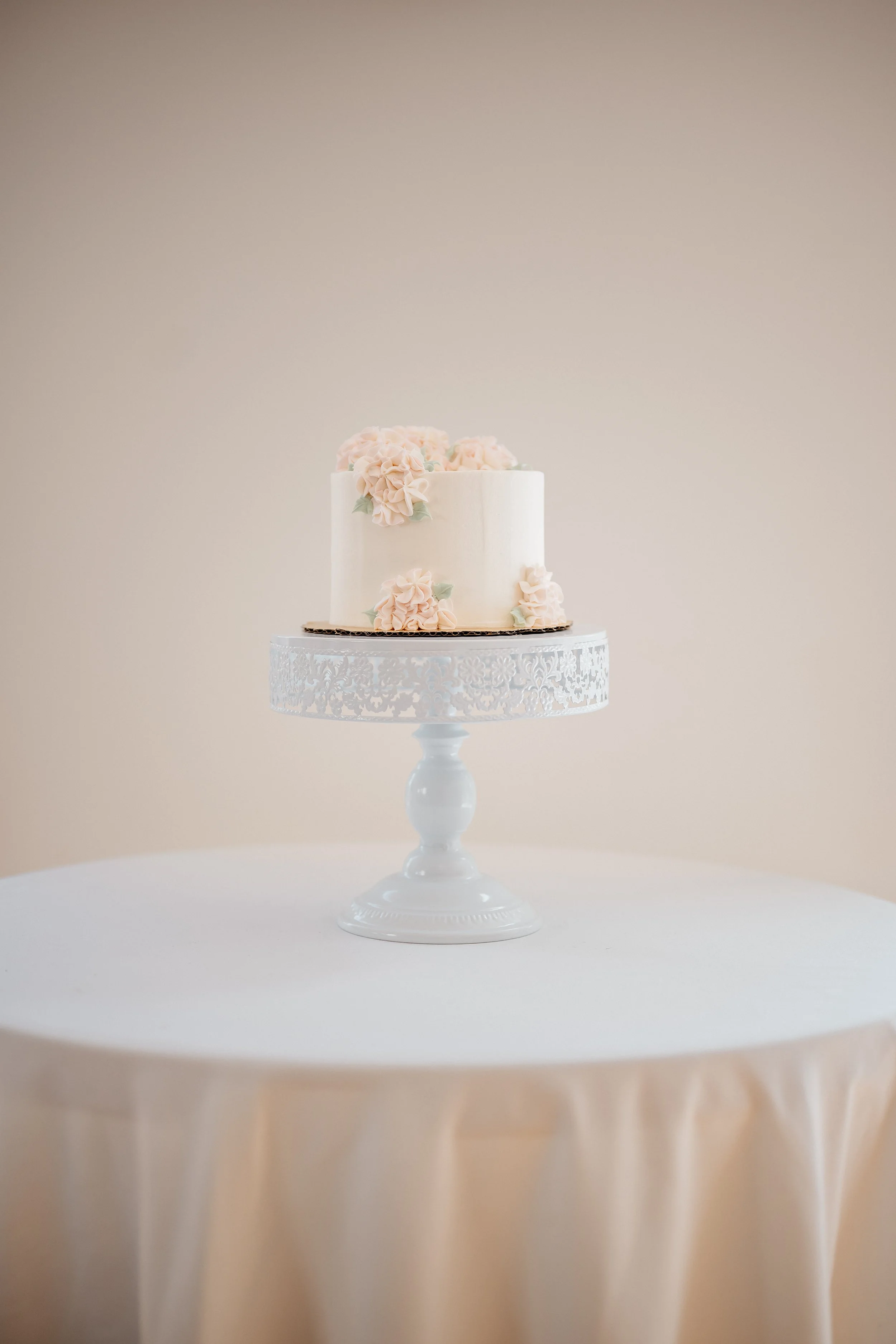 A small white wedding cake with pink flowers on a white ornate cake stand, placed on a round table with a white tablecloth.