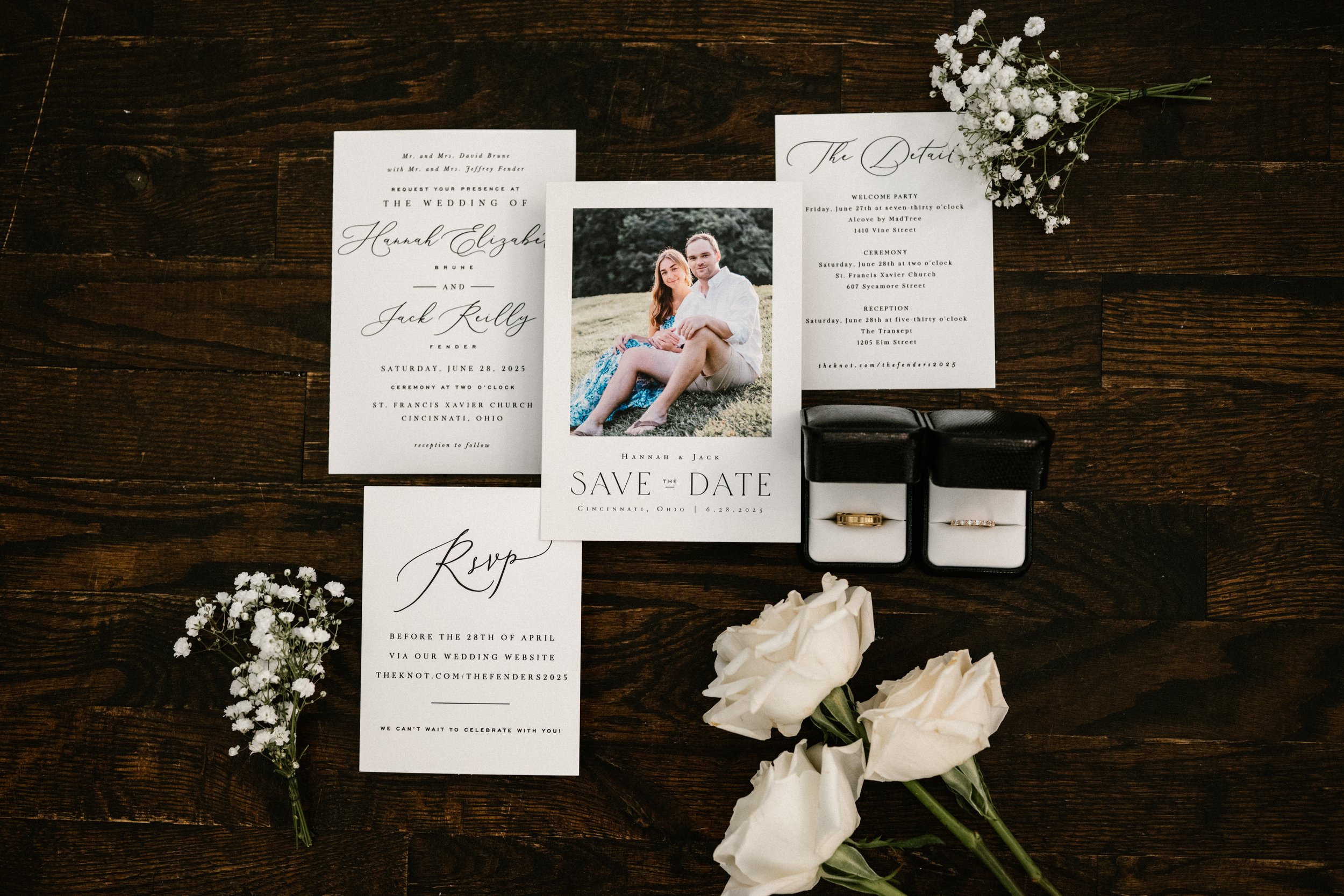 Cincinnati Ohio Wedding invitation suite with a photo of a couple, wedding rings in boxes, white roses, baby's breath flowers, and a bouquet of white roses, all arranged on a dark wooden surface. wedding day flat lay.