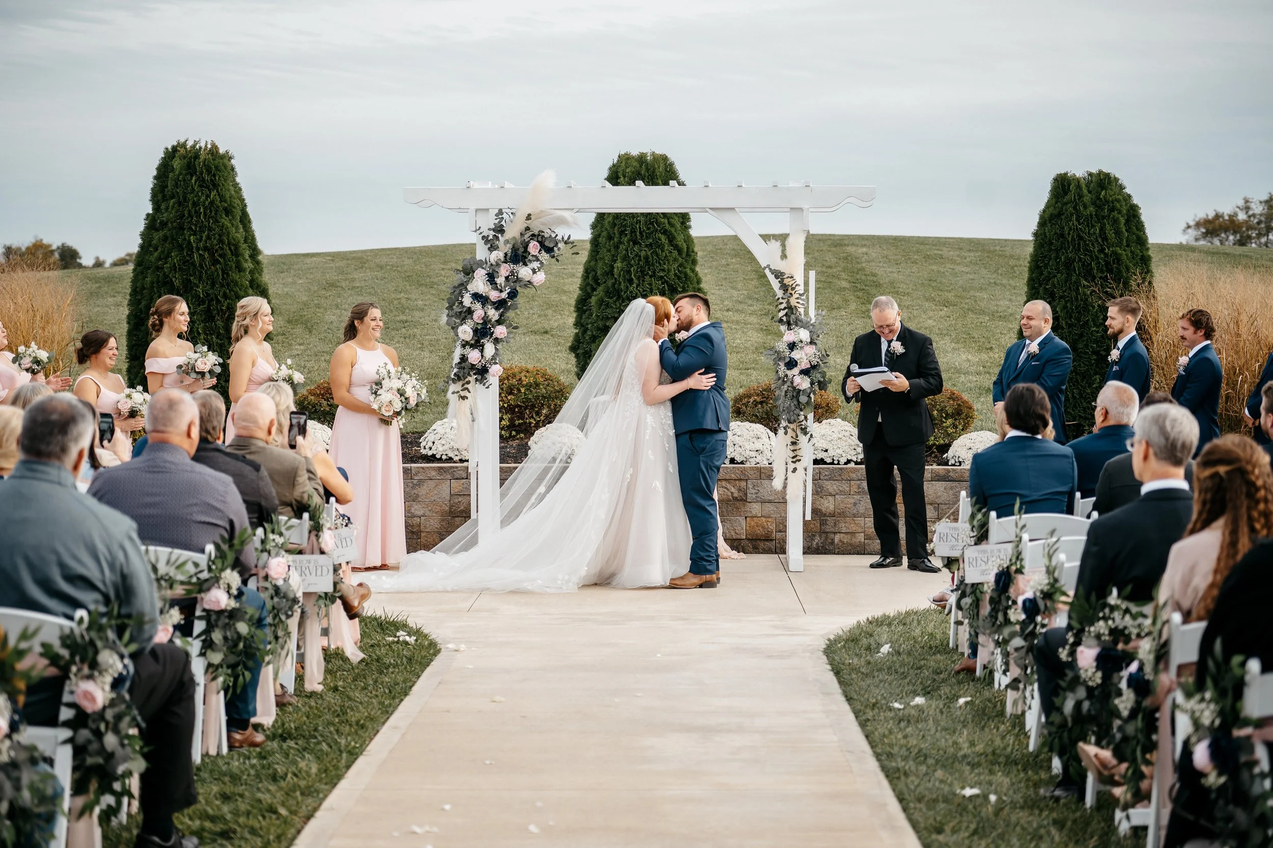 A Dayton Ohio wedding ceremony outdoors with a bride and groom first kiss under a decorated arch, surrounded by guests seated on either side.