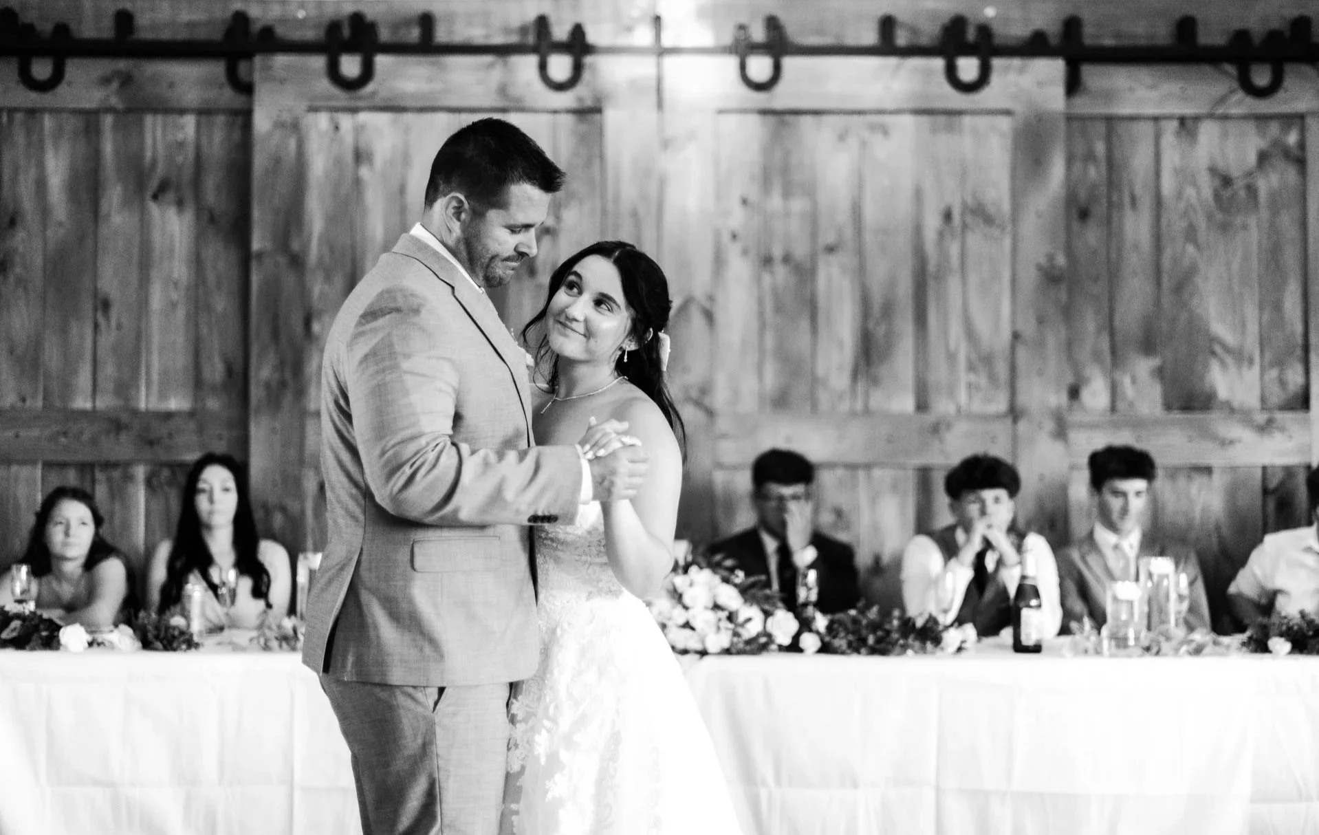 A bride and her father slow dancing at their wedding reception, with guests seated at a head table decorated with flowers in the background.