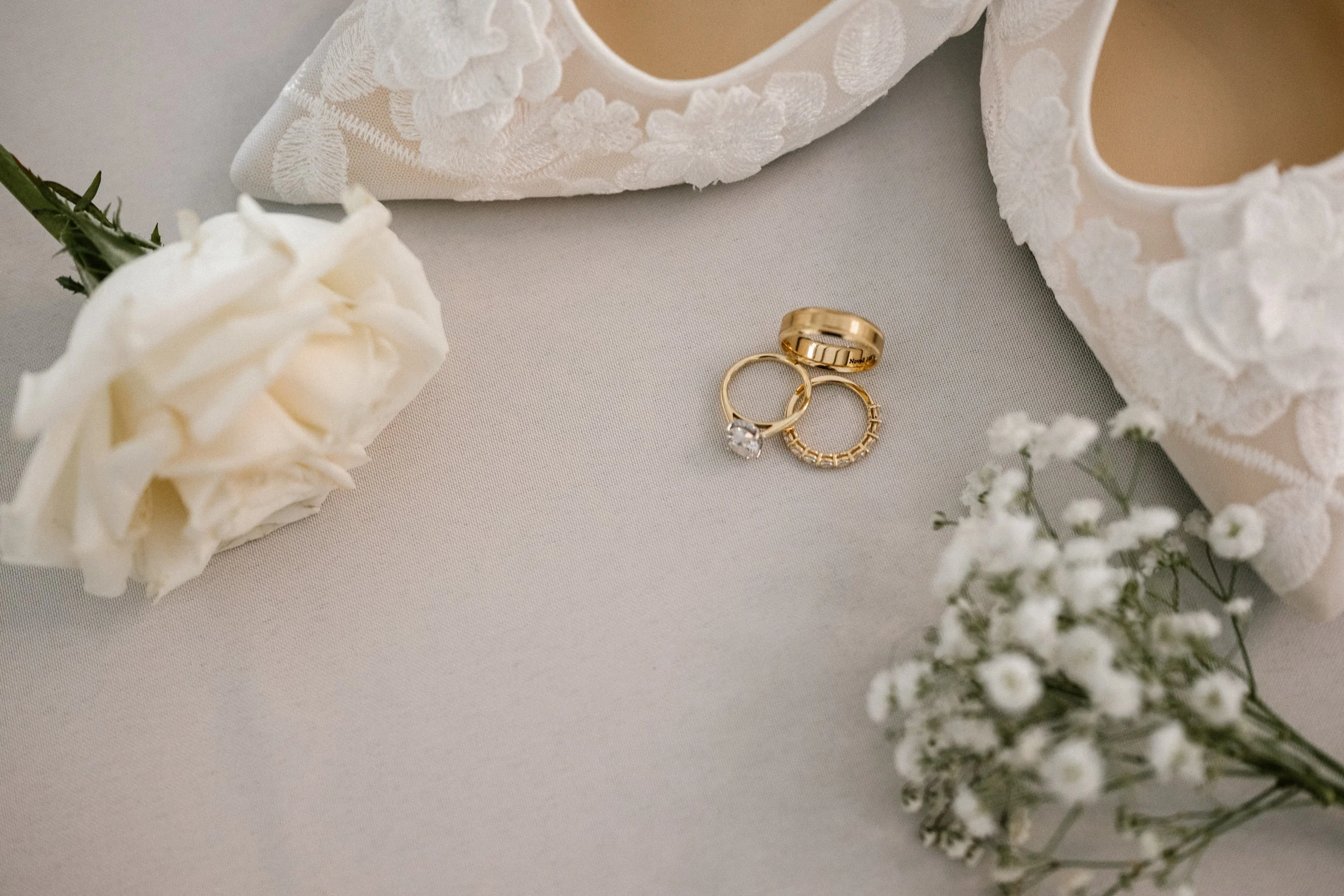 Three gold wedding rings, one with a diamond, placed on a light-colored surface with white roses and a pair of white lace shoes nearby.