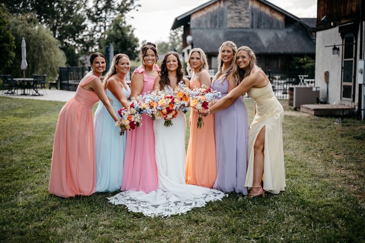 bride with bridesmaids in Dayton Ohio wearing colorful dresses holding bouquets, standing on grass in front of a rustic barn during daytime.