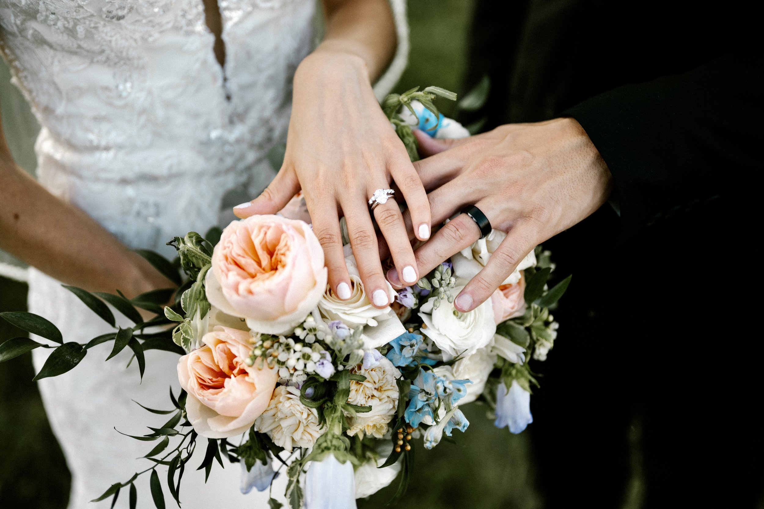 Close-up of a Cincinnati Ohio bride and groom's hands showing wedding rings, resting on a bouquet of flowers with peach, white, and blue blossoms, and greenery.