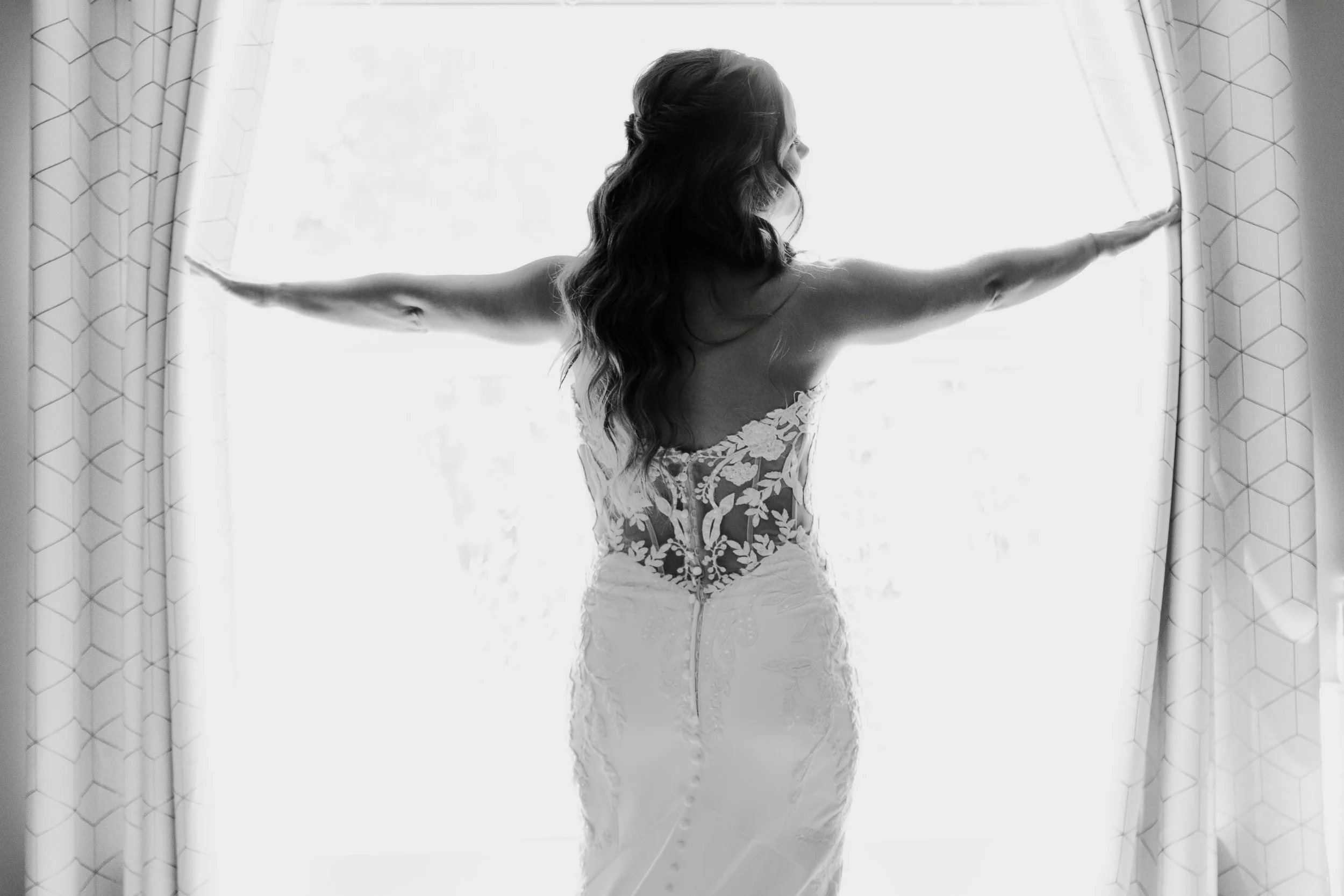 A Dayton Ohio bride standing infant of a window pulling the curtains open in black and white showing the back of the dress in detail.
