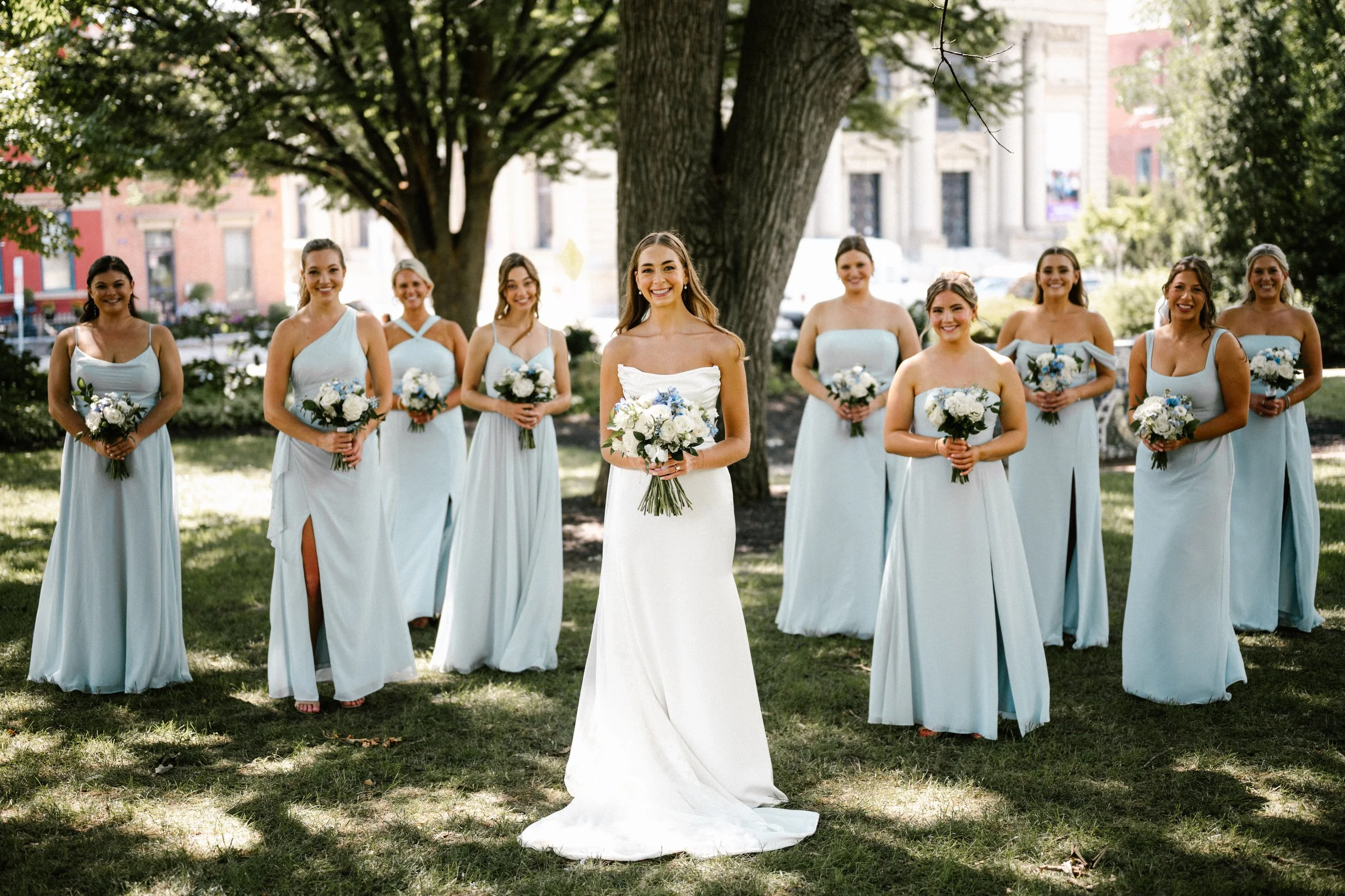 A Cincinnati Ohio bride in a white wedding dress standing with her bridesmaids in light blue dresses holding bouquets of white and blue flowers outdoors under a large tree on a sunny day.
