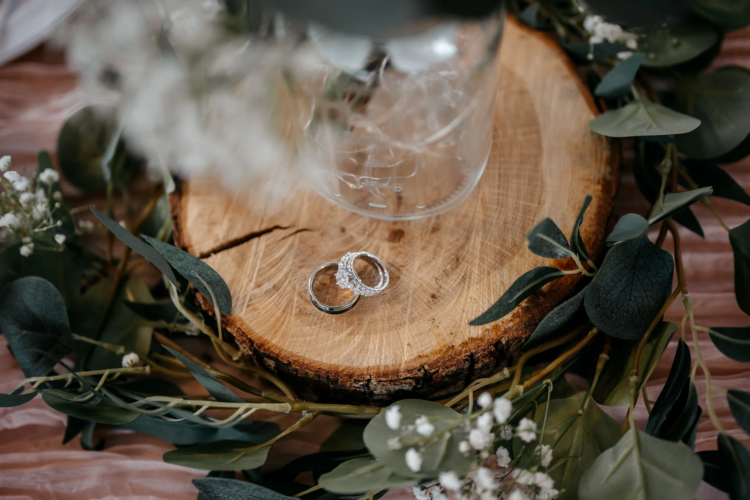 A wooden slice on a pink fabric surface, decorated with green leaves and small white flowers, holds a clear glass container. Two wedding rings, one with diamonds and the other plain, are placed on the wood in Dayton Ohio.