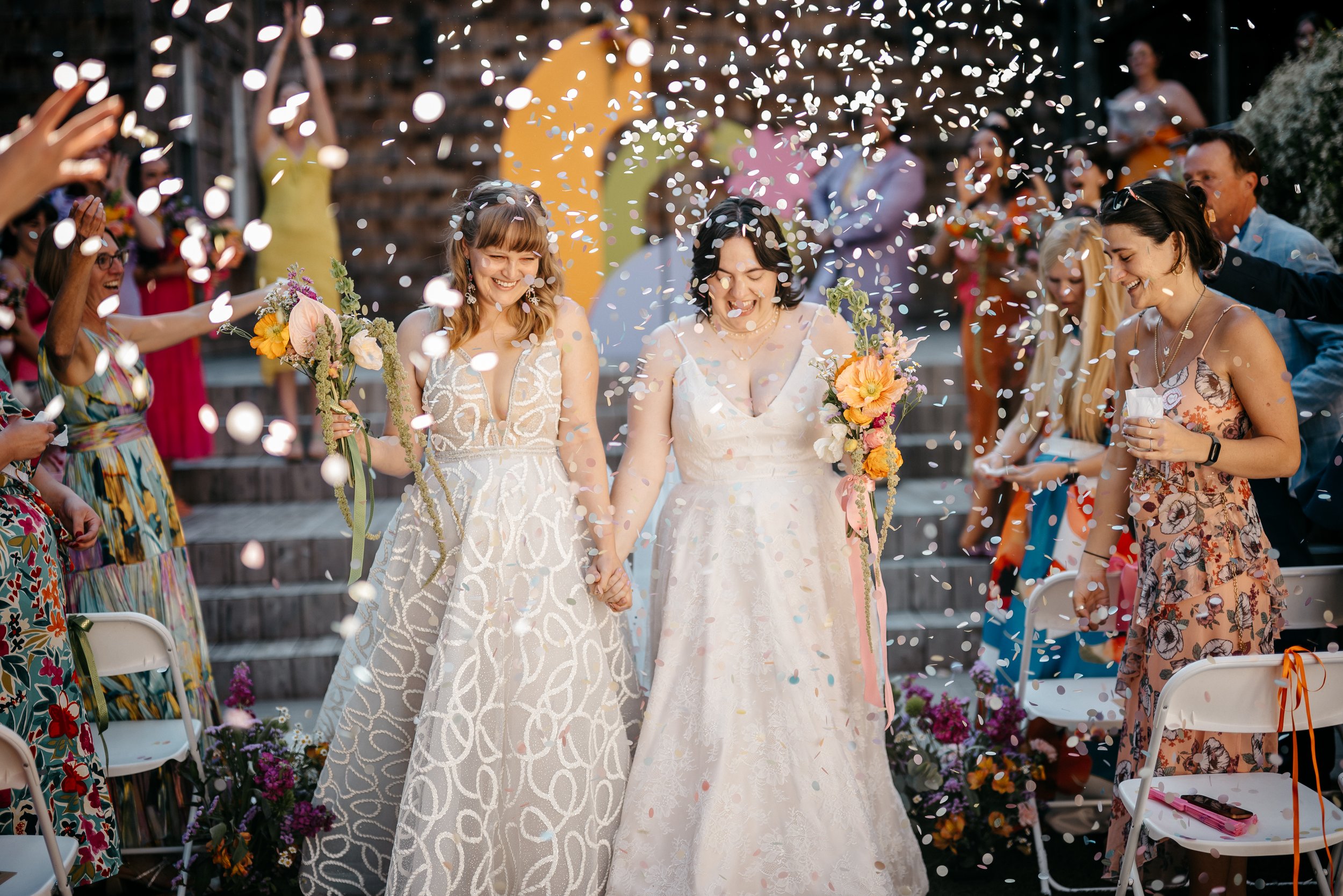 Two Cincinnati Ohio brides holding hands and smiling as they walk through falling confetti at a wedding celebration, surrounded by guests and colorful decorations.