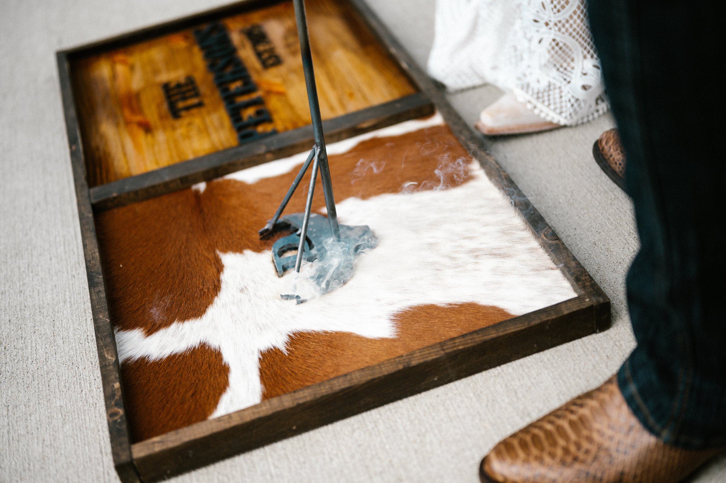 A Dayton Ohio newlywed bride and groom is using a branding iron on a piece of cowhide, which is inside a wooden box on the ground. The person's right foot is visible, wearing a brown cowboy boot.