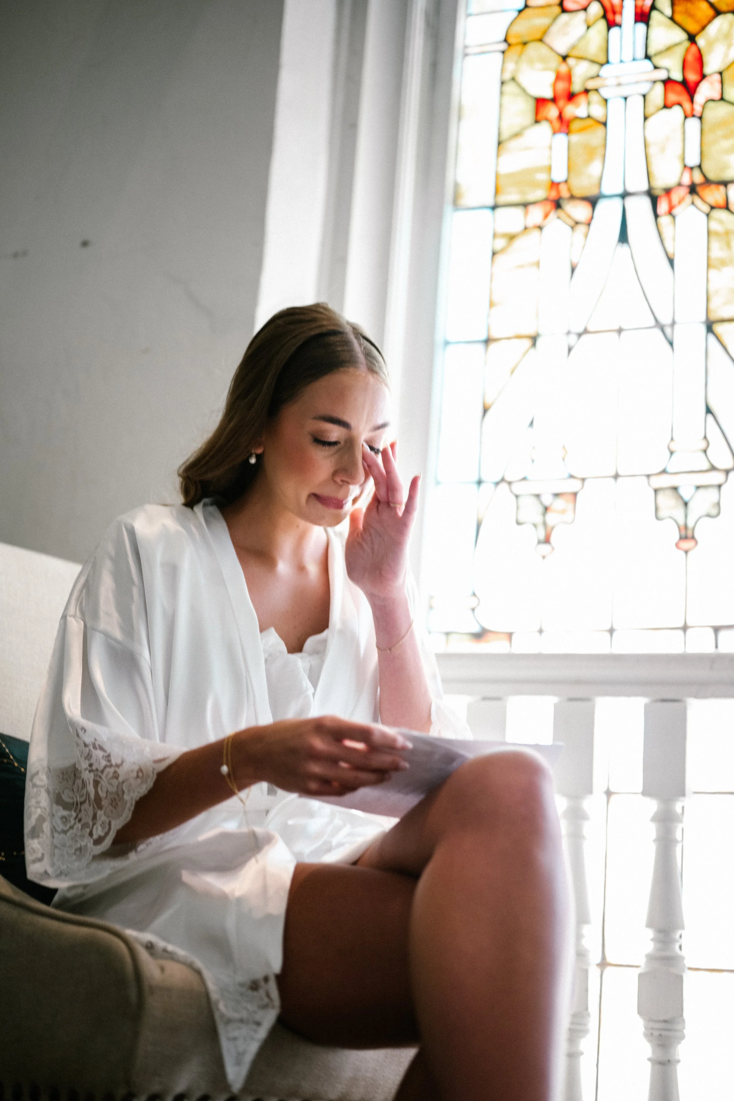 A Cincinnati Ohio bride in white, sitting on a beige chair near a stained glass window, appears emotional, with one hand touching her face and the other holding a note from groom on her wedding day.