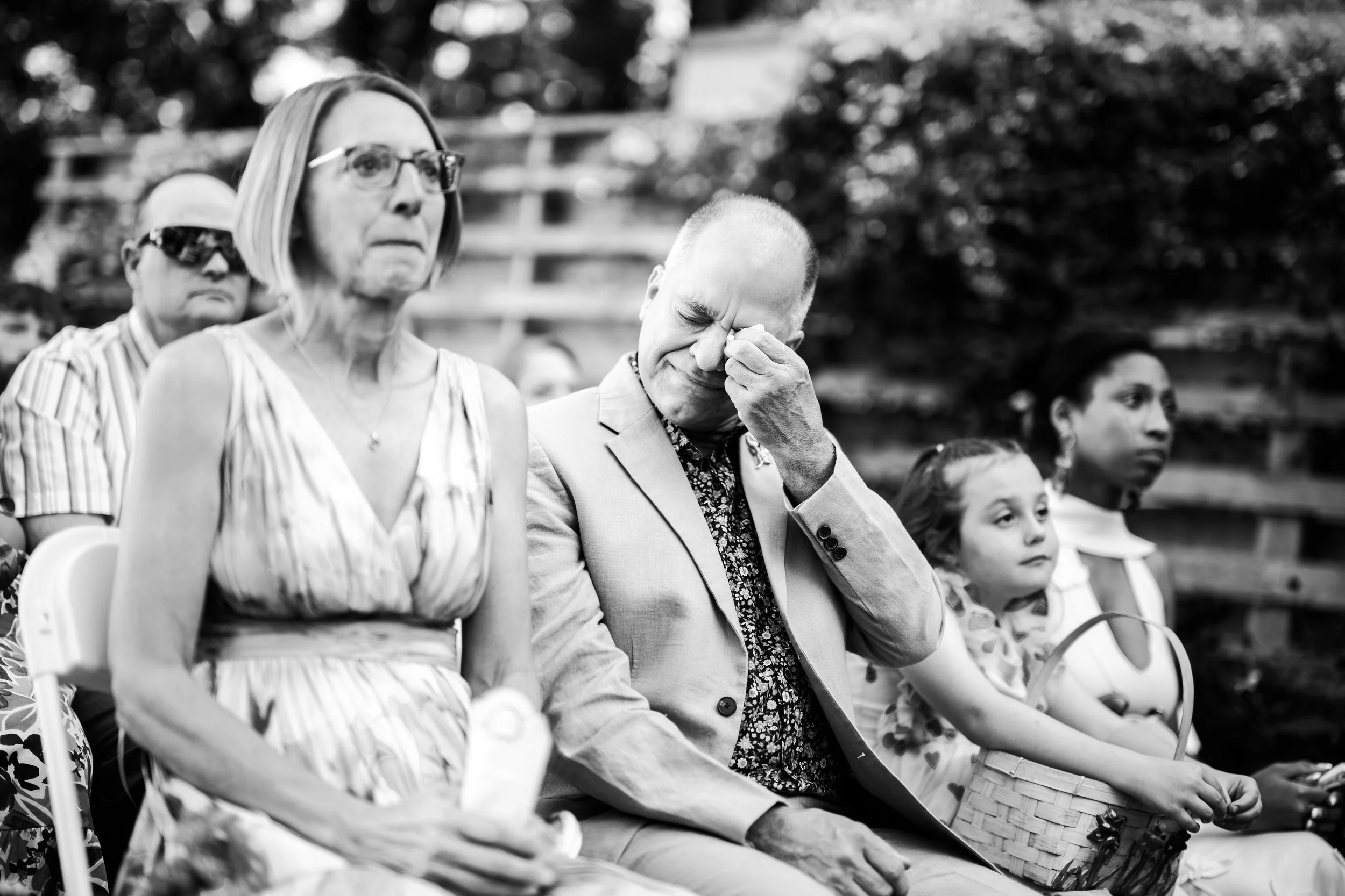 People sitting outdoors, some appearing emotional; a father of the bride and mother of the bride in the center crying, wiping his eye, others showing somber expressions.