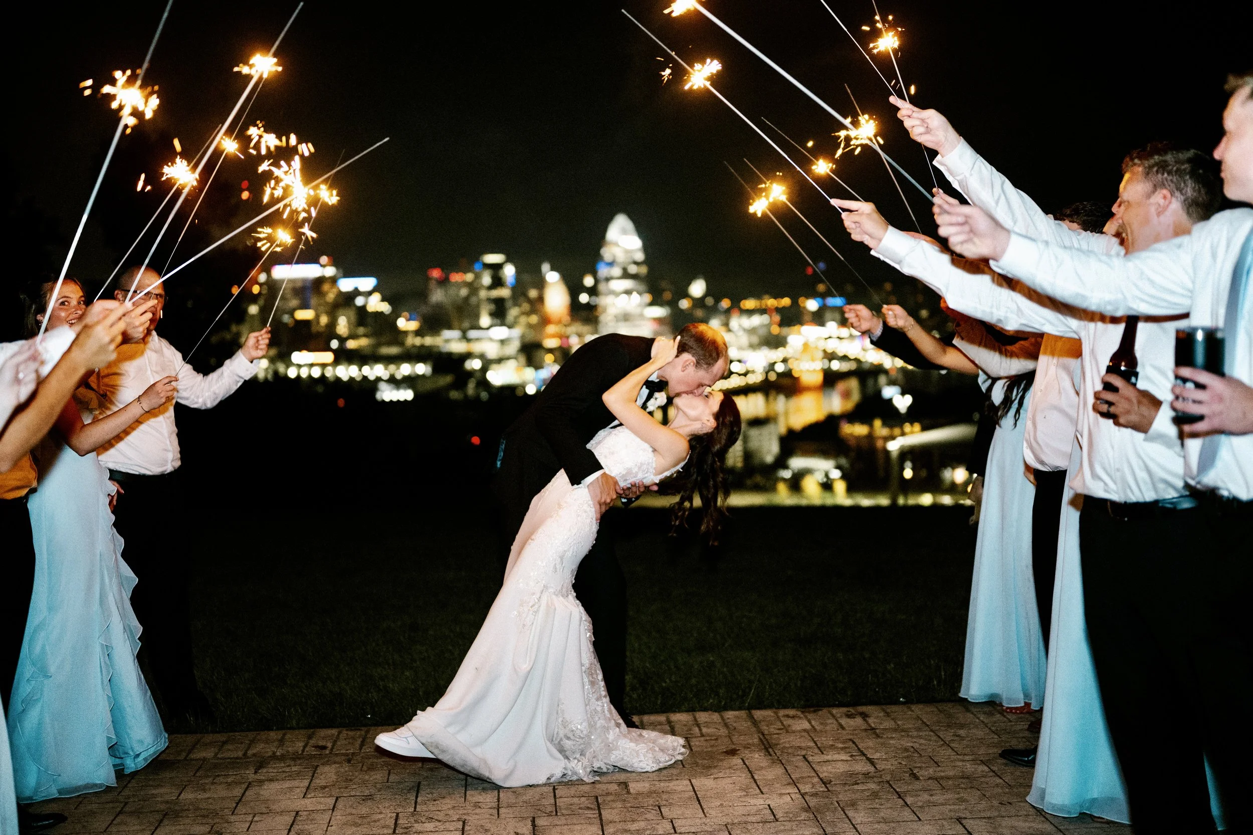Wedding celebration at night in Cincinnati Ohio with the bride and groom kissing while surrounded by friends holding sparklers; city skyline with illuminated buildings in the background.