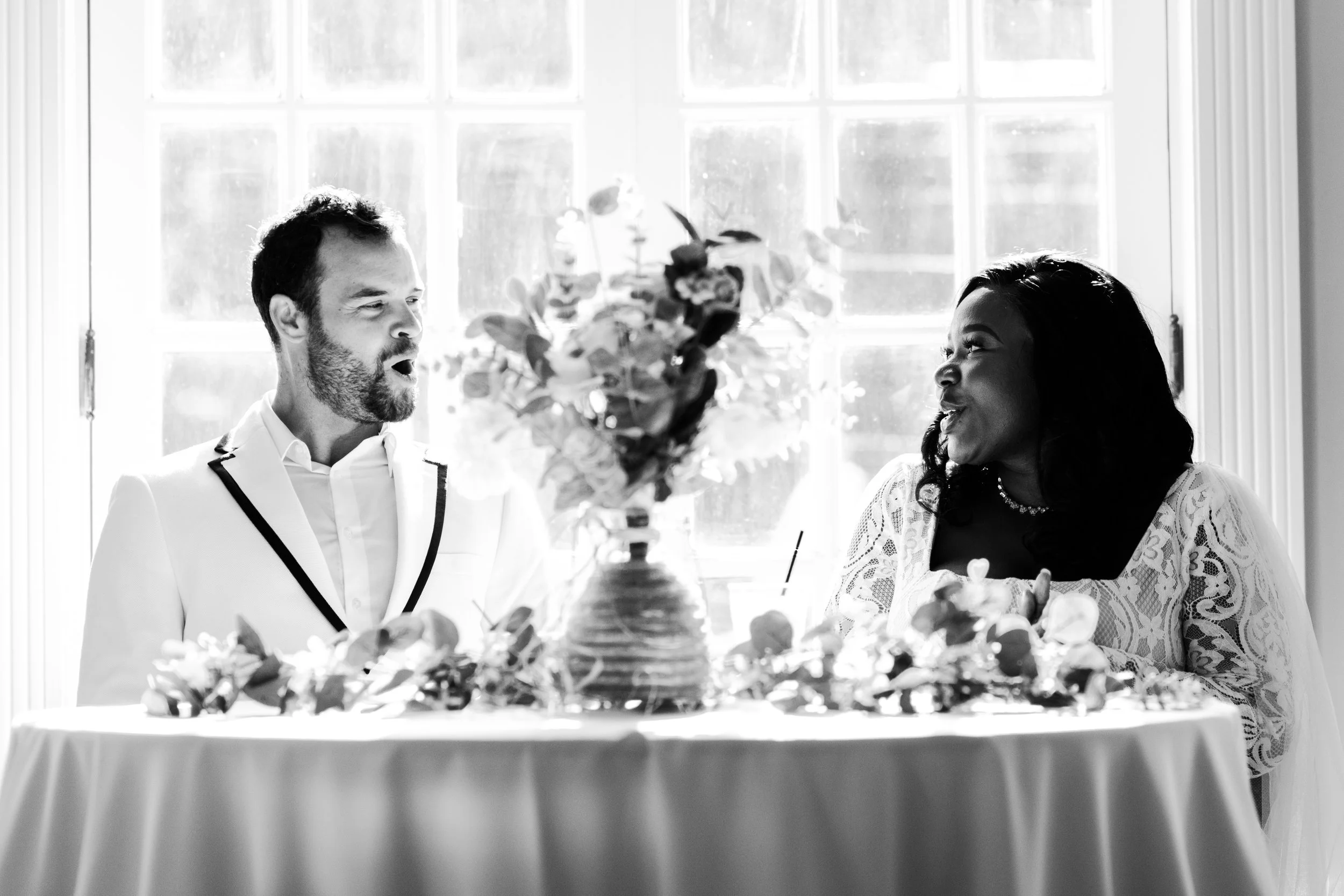 Black and white photo of a couple sitting at a table with floral decorations, engaged in conversation in front of a window.