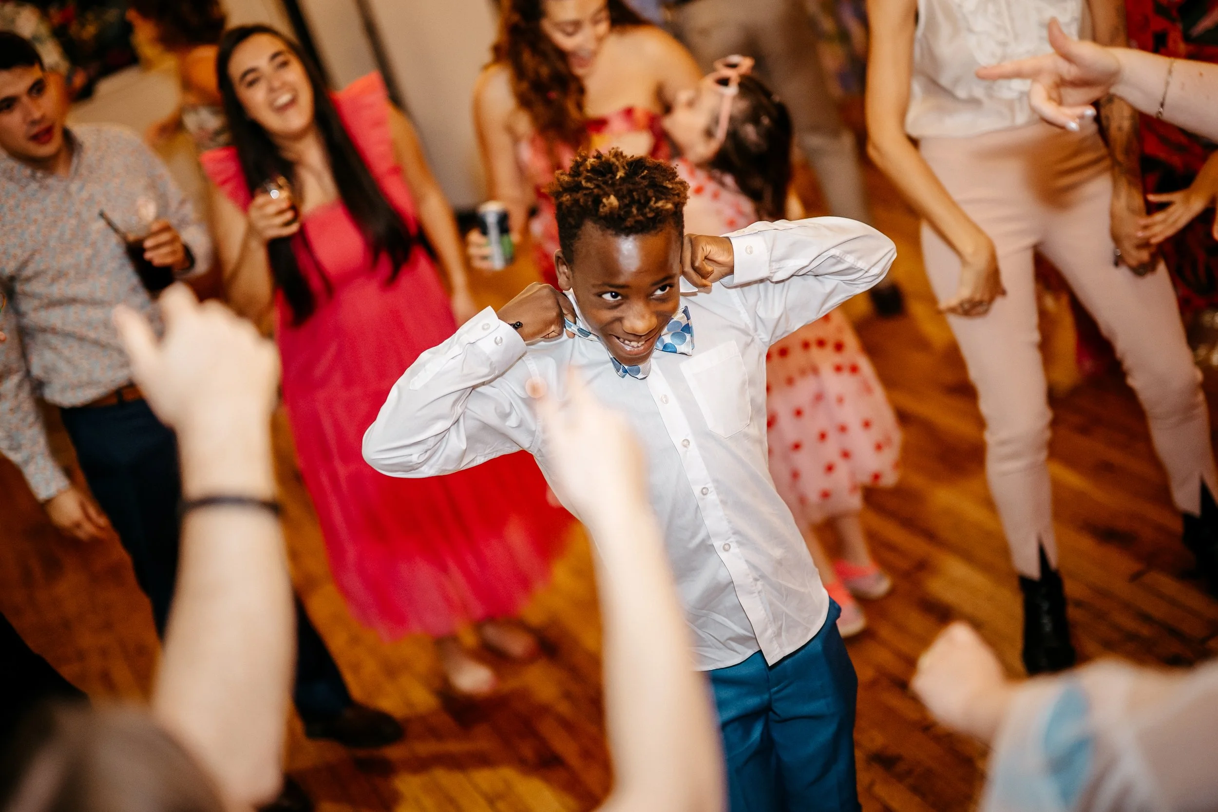 A Cincinnati Ohio boy dancing and smiling at a lively wedding party surrounded by adults and children, some holding drinks, in a warmly lit indoor space.