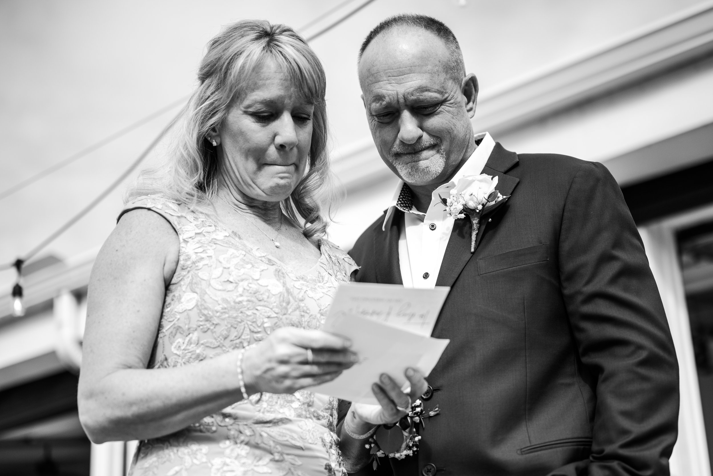 A Dayton Ohio brides mother in a lace dress and a brides father in a suit with a boutonniere read a brides note together, both showing emotional expressions, at a formal event.
