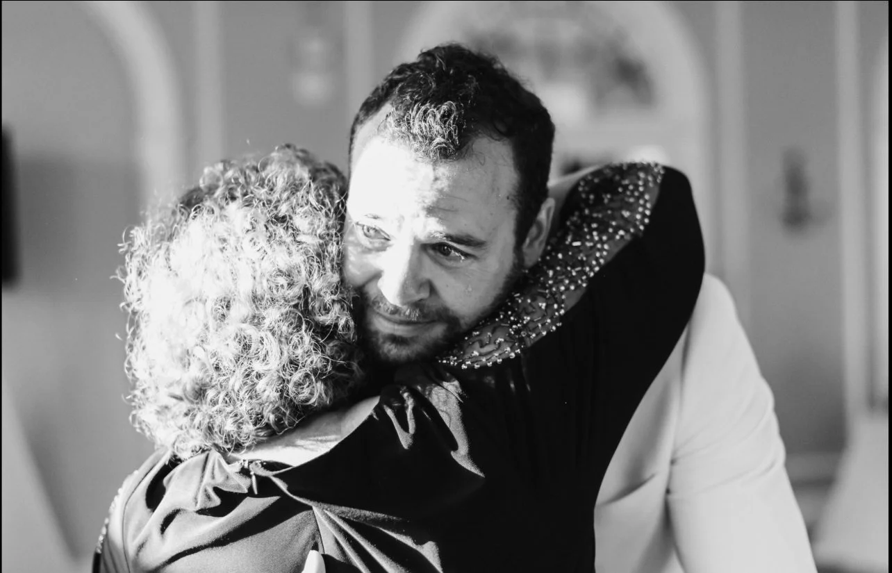 A Dayton Ohio groom with short dark hair and a beard hugging his mother, crying, with curly hair in a black jacket with a sequin collar.