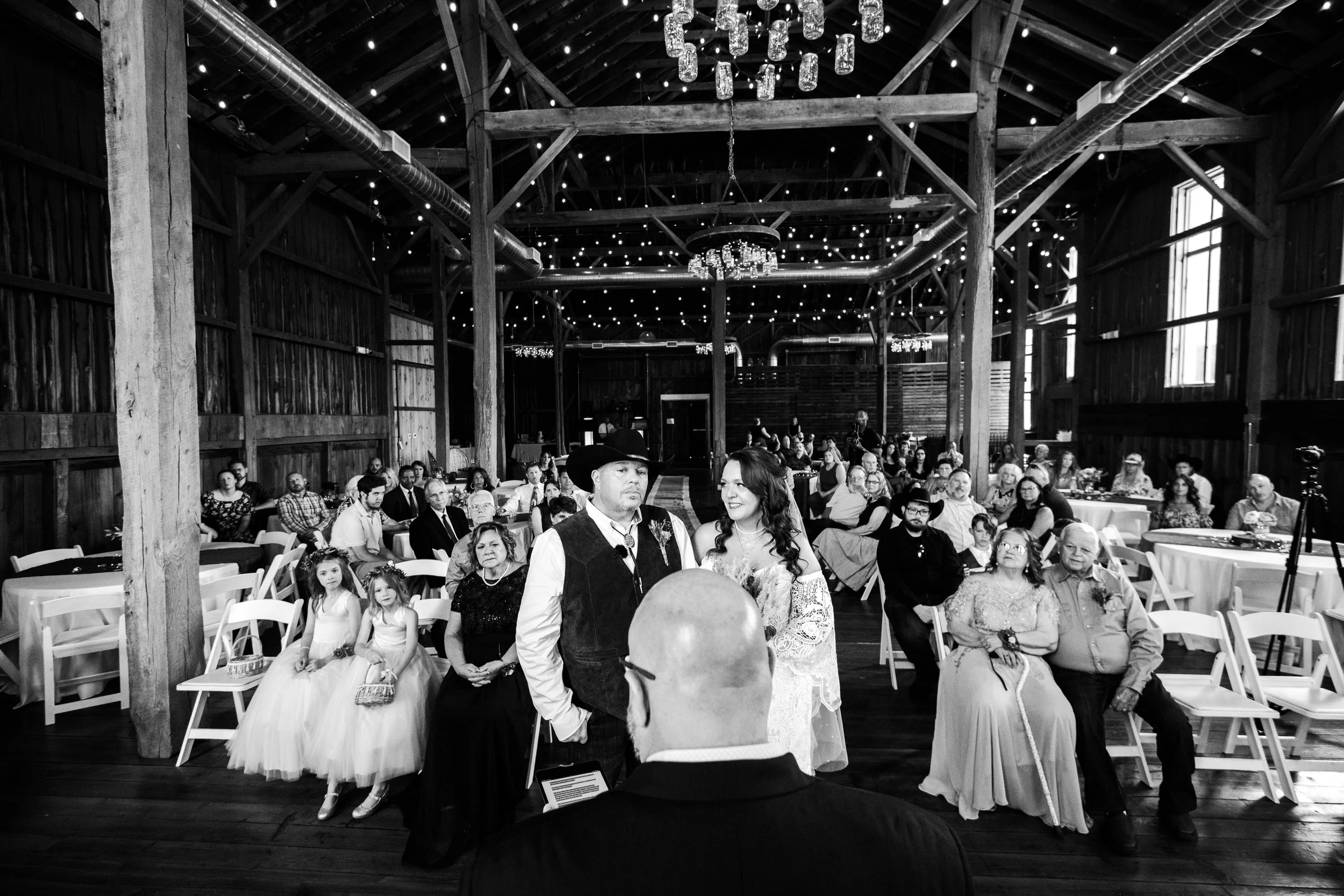A Dayton Ohio wedding ceremony inside a rustic barn with wooden beams and string lights, featuring a bride and groom, officiant, and seated guests.