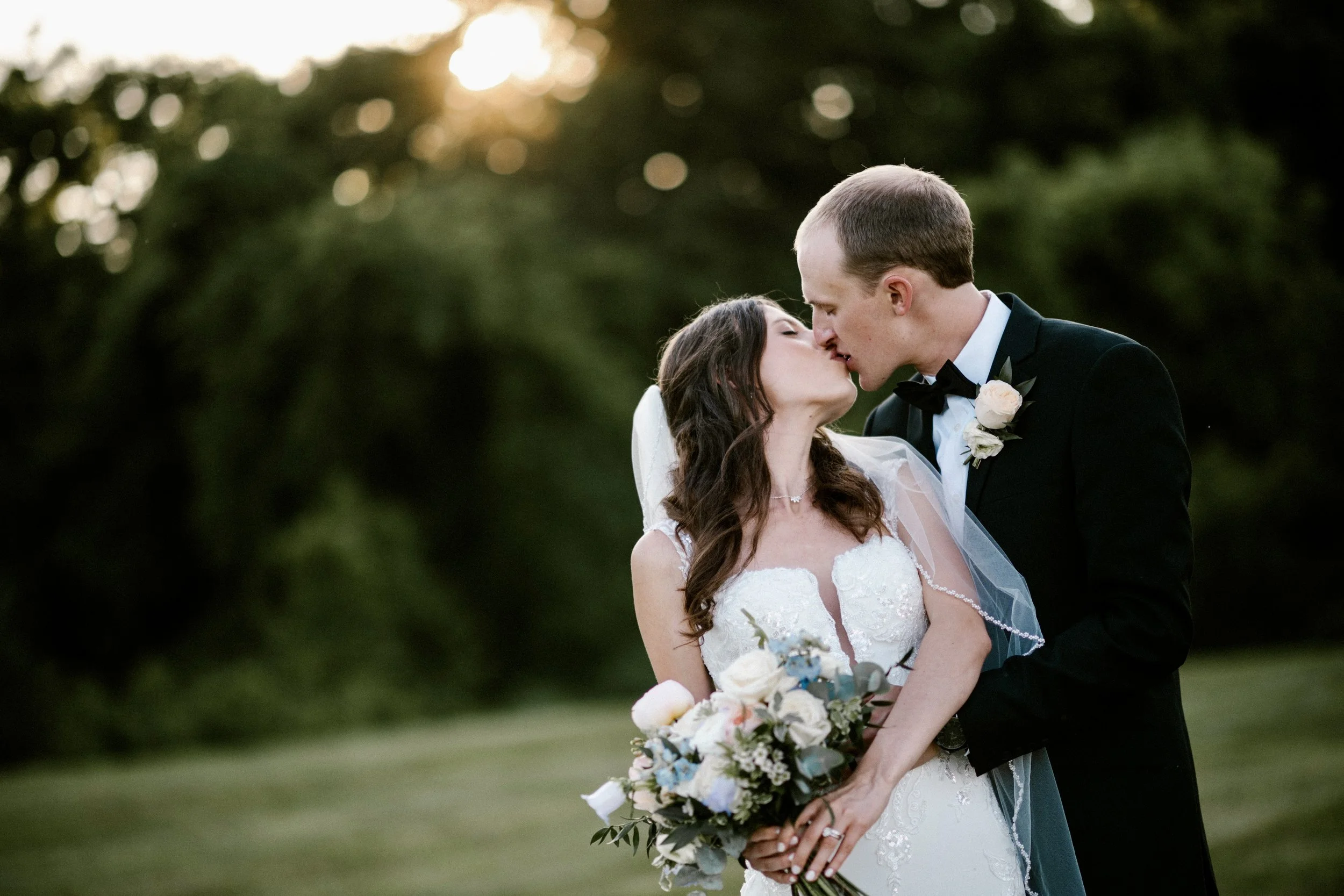 A bride and groom share a kiss outdoors during sunset, with trees in the background. The bride is holding a bouquet of flowers, and the groom is dressed in a tuxedo with a boutonnière.