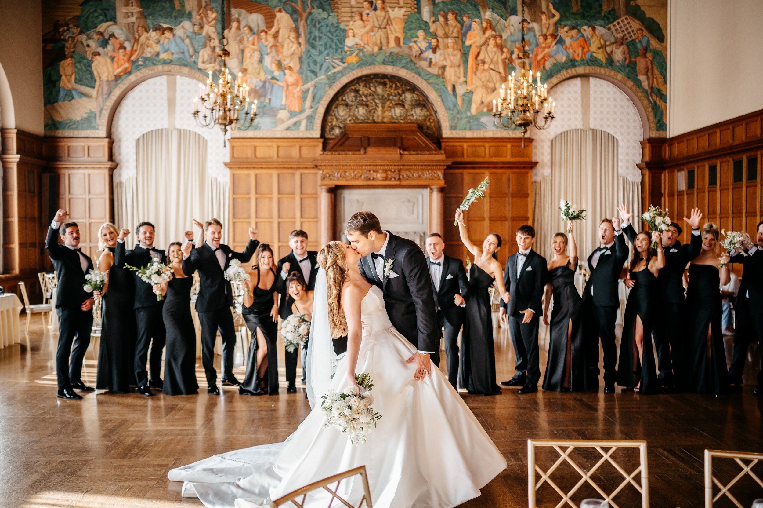 A Cincinnati ohio wedding reception with a bride and groom kissing in the center, surrounded by cheering wedding party holding bouquets, in an elegant hall with wood-paneled walls, chandelier lighting, and a large ornate mural.