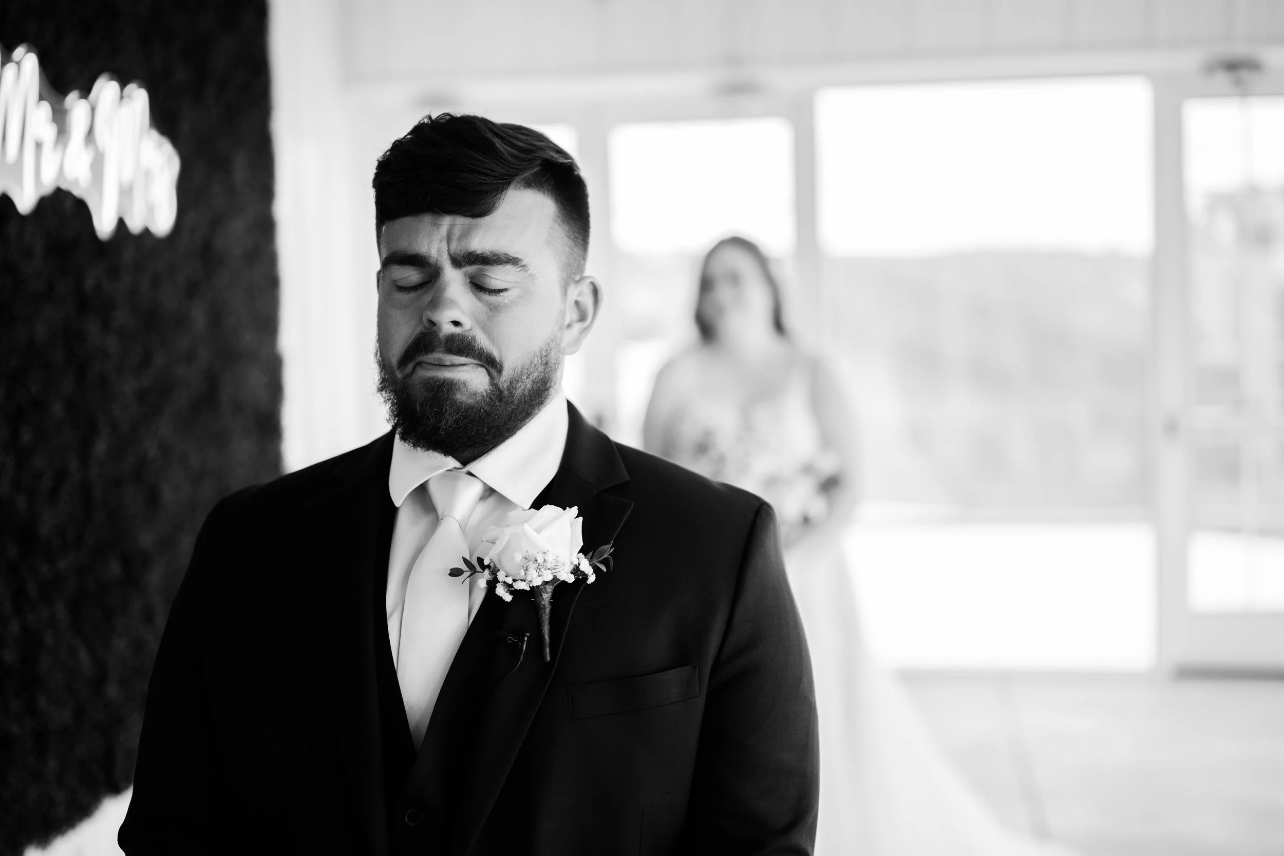A Dayton Ohio groom with a beard dressed in a tuxedo and a boutonniere is standing with eyes closed, appearing emotional, waiting to do first look with his bride in a room with large windows. A bride in a white dress is seen blurred in the background
