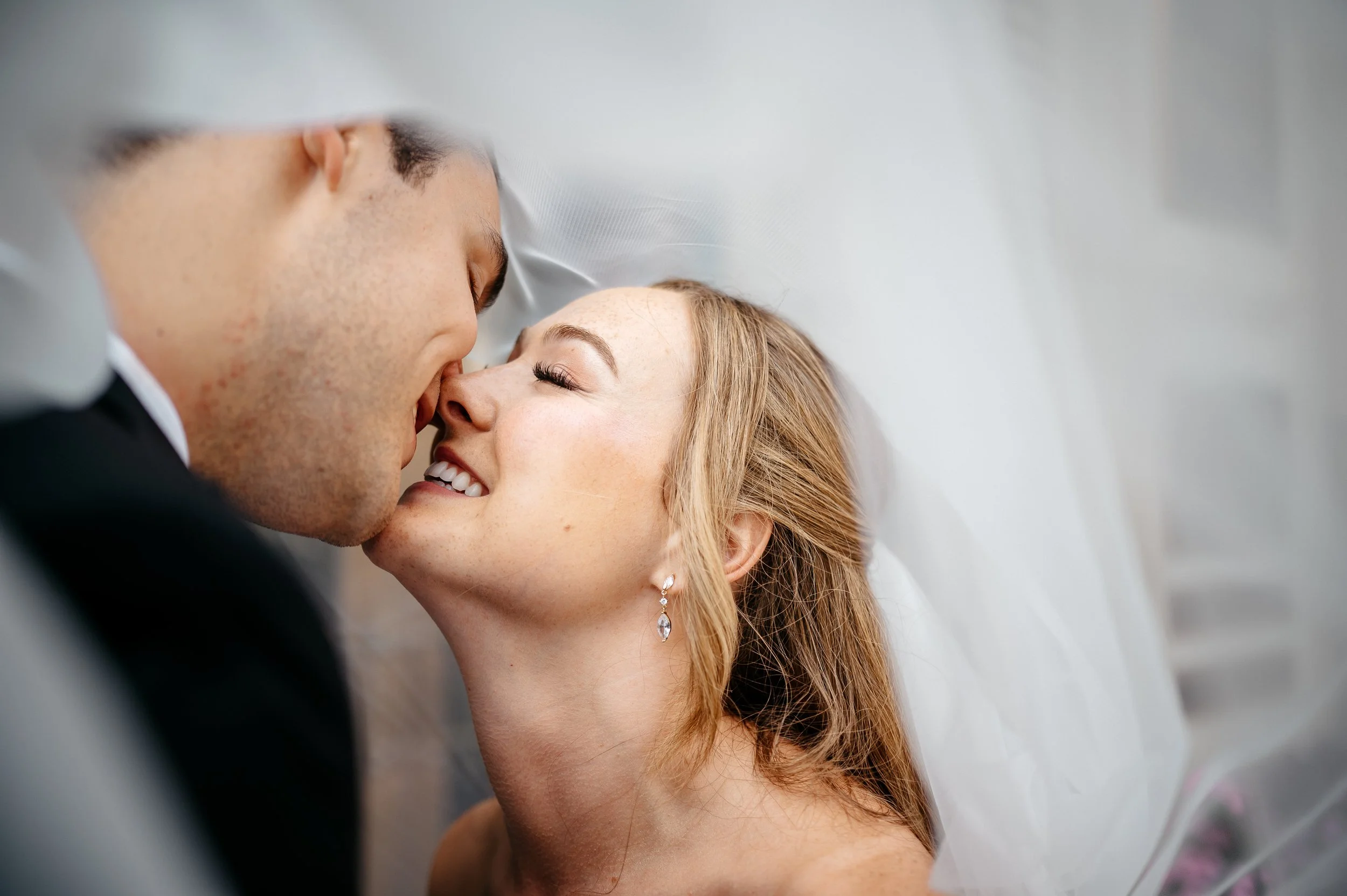 A bride and groom share a close, intimate moment with their lips touching, eyes closed, smiling in a wedding setting.