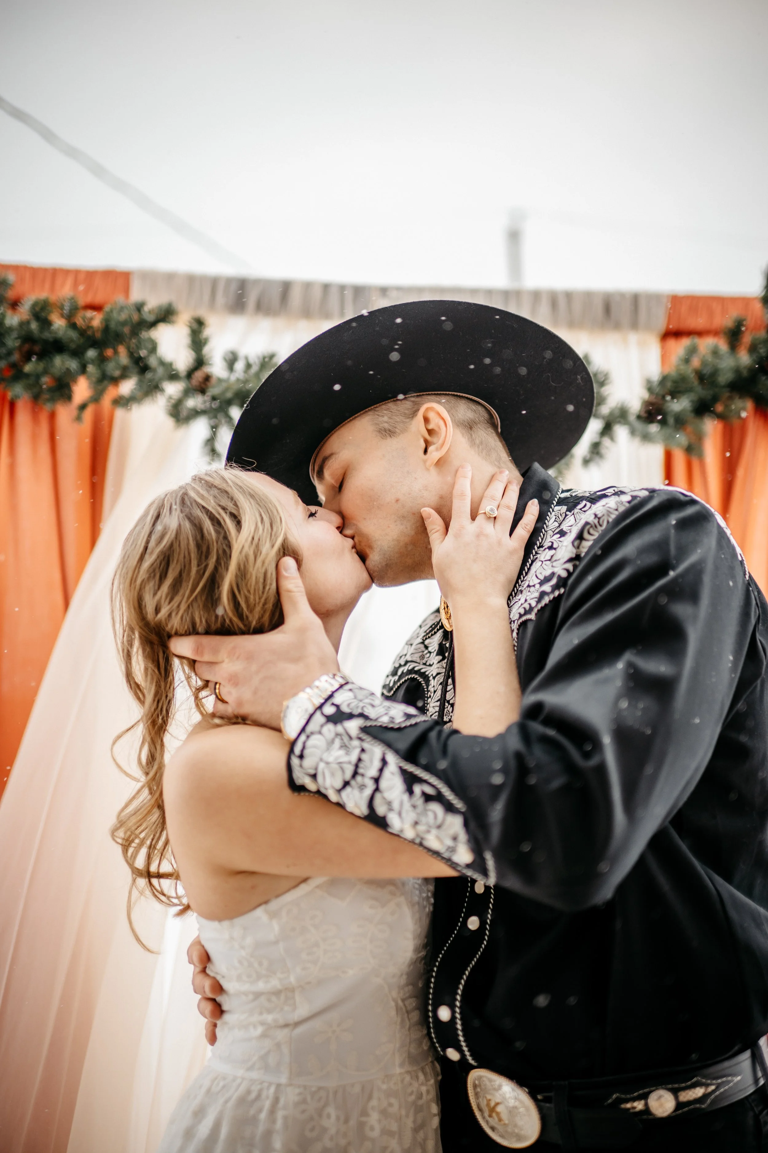 A Dayton Ohio bride and groom sharing a kiss during wedding, with the woman in a white dress and the man in traditional Texas cowboy attire, including a black cowboy hat. They are embracing in front of a decorated backdrop with greenery and orange cu
