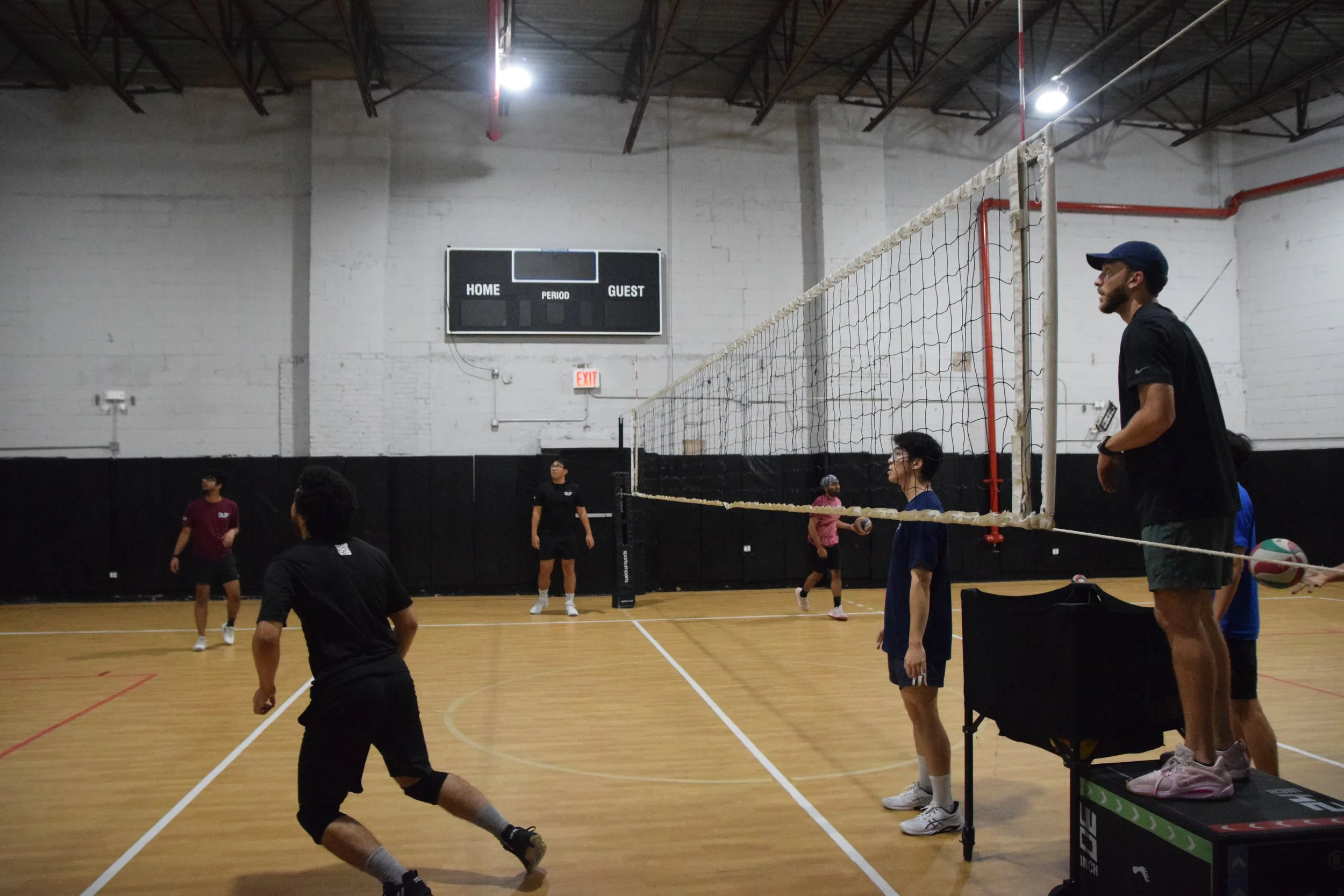 Young people playing indoor volleyball in a gym. Two players are near the net, one on the stand and one preparing to hit the ball, while others observe or prepare to play. The gym has a wooden floor and a scoreboard on the wall.