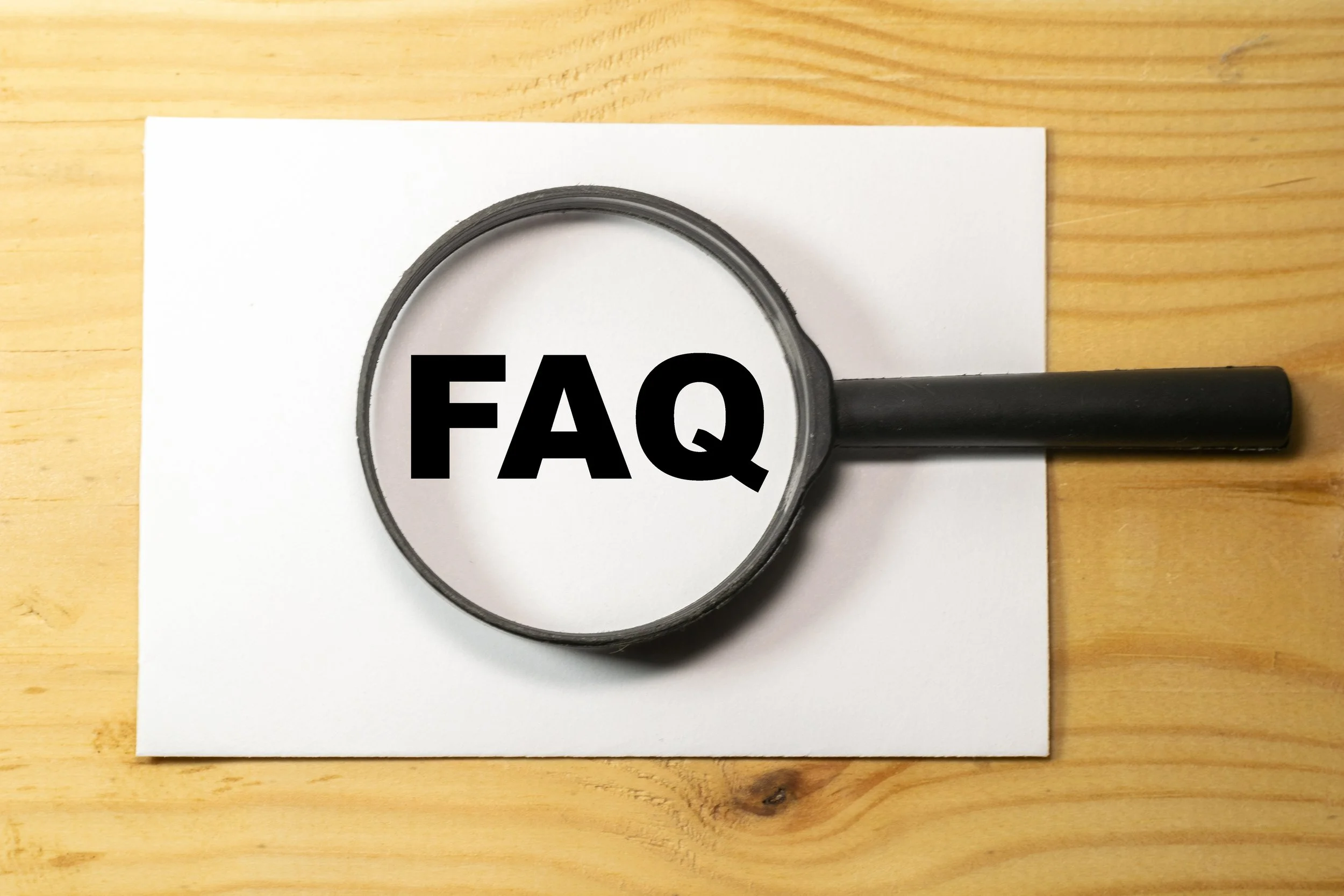 Magnifying glass with the text FAQ on a white paper on a wooden surface.