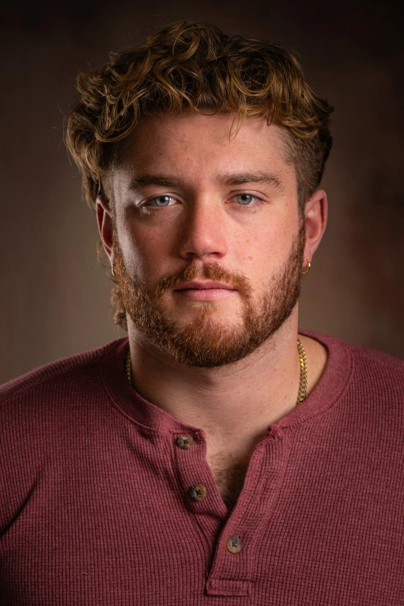 Close-up, three-point lighting, portrait of a young man with curly light brown hair, blue eyes, and a full beard and mustache, wearing a maroon button-up shirt with a gold chain necklace.