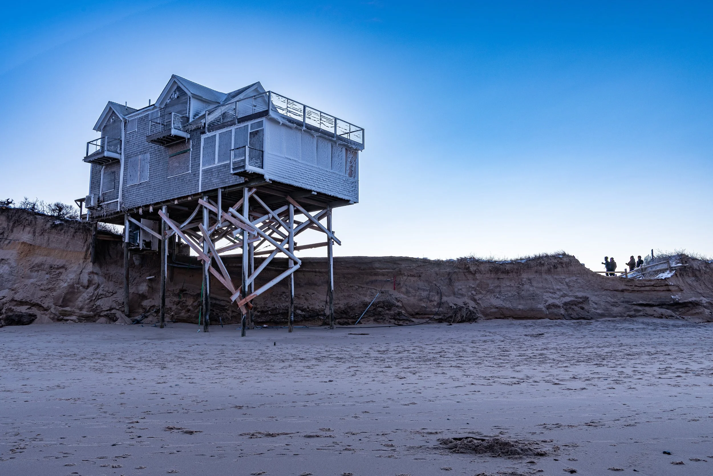 A house on stilts atop a sandy, eroded cliff with a clear blue sky in the background. Four small figures are seen in the distance on the right.