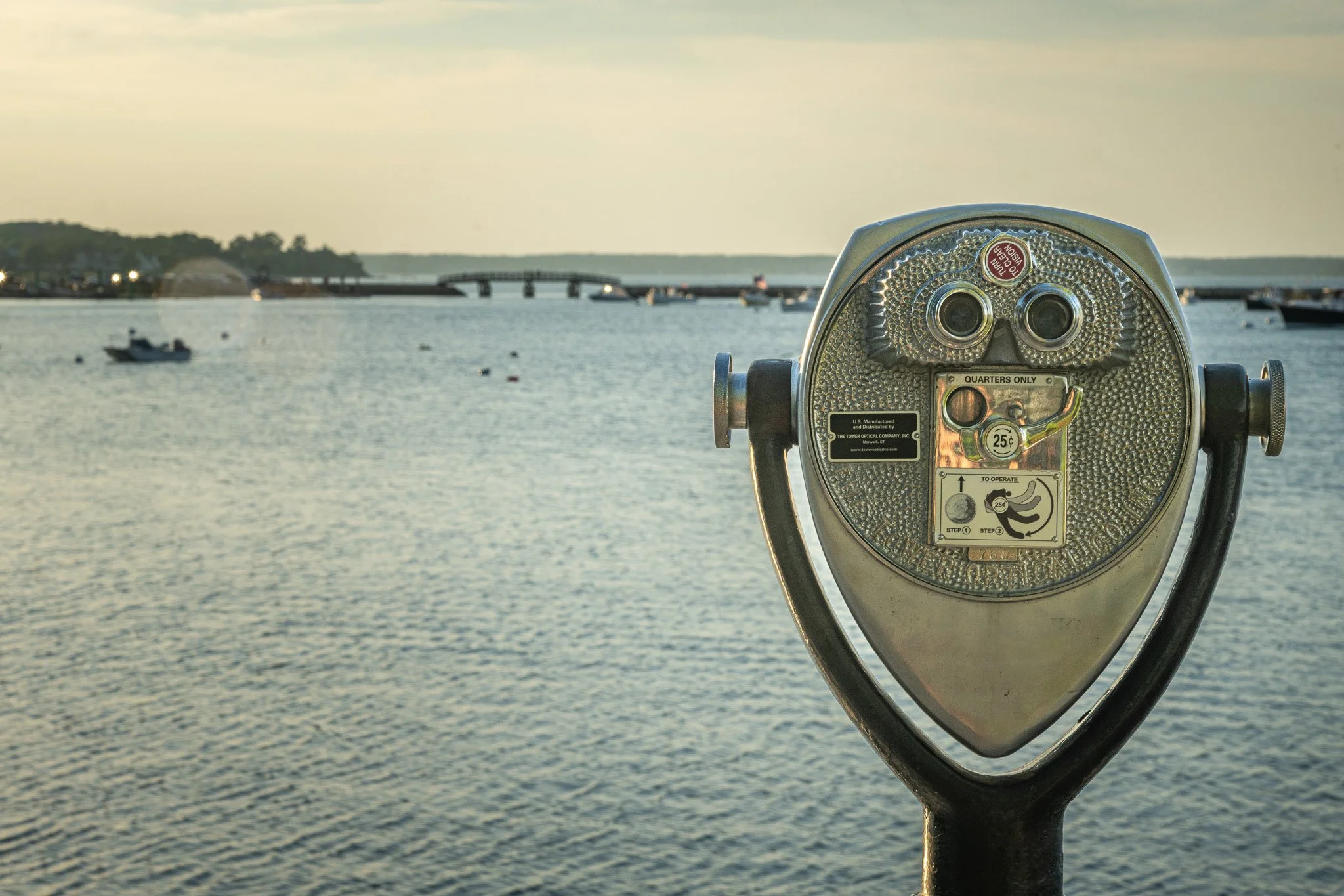 A coin-operated binocular viewer facing a body of water with a distant shoreline, boats, and a bridge, during sunset or late afternoon.
