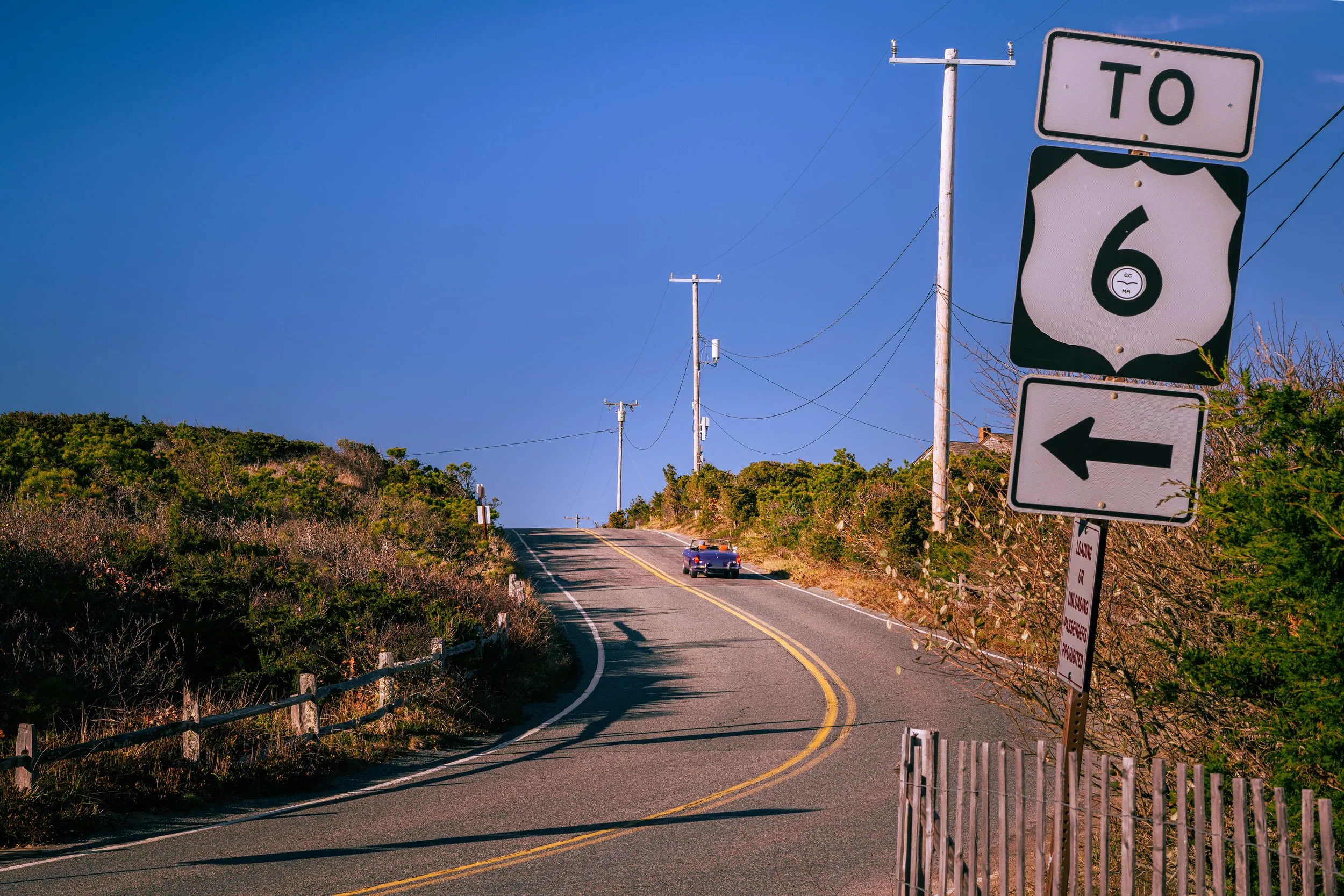 The image shows a rural road with a slight bend, a car driving away in the distance, utility poles and wires along the side of the road, and multiple road signs including a shield-shaped sign indicating U.S. Route 6 and a rectangular sign with a left arrow. There are bushes and trees on either side of the road under a clear blue sky.