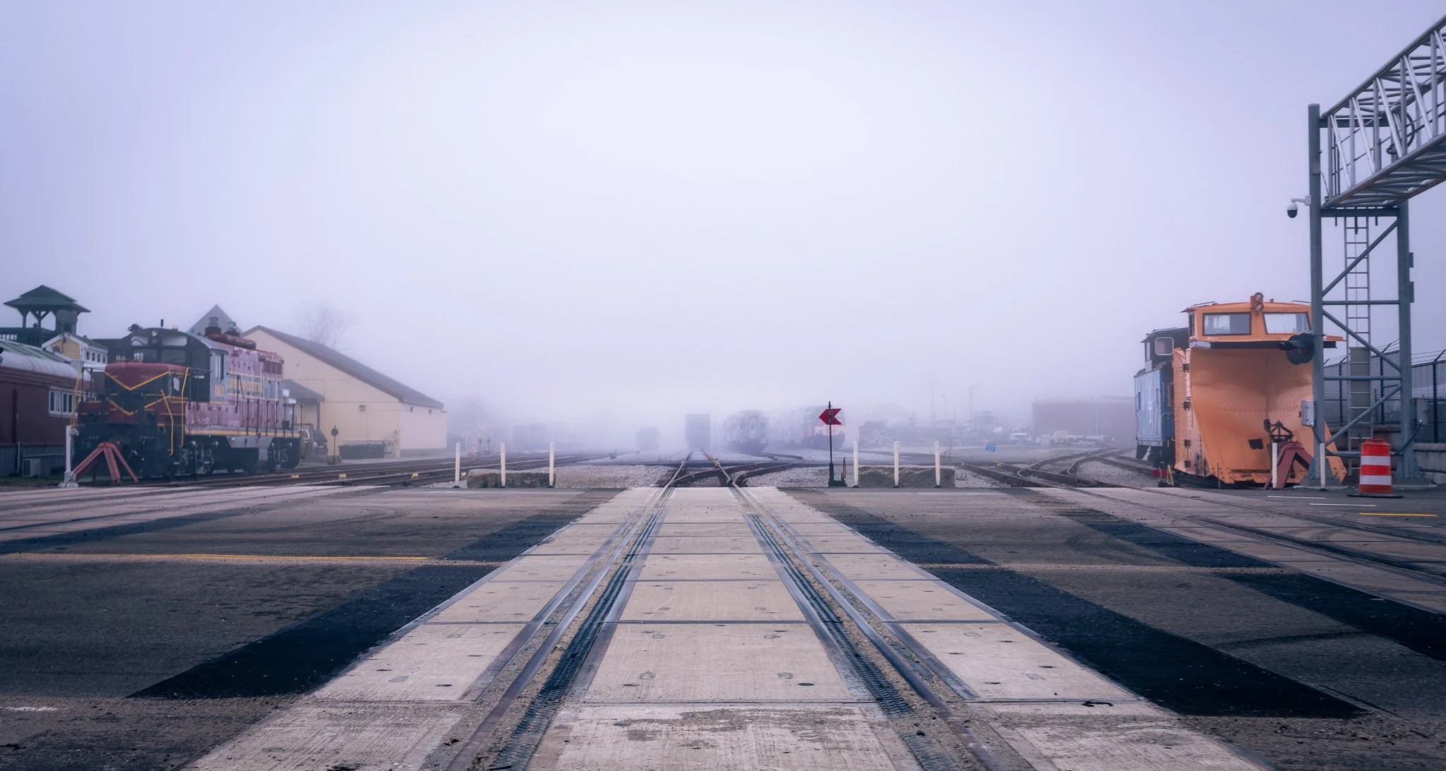 A foggy railroad yard with train tracks, two locomotives, and a small train station building in the background, along with railway equipment and a platform.