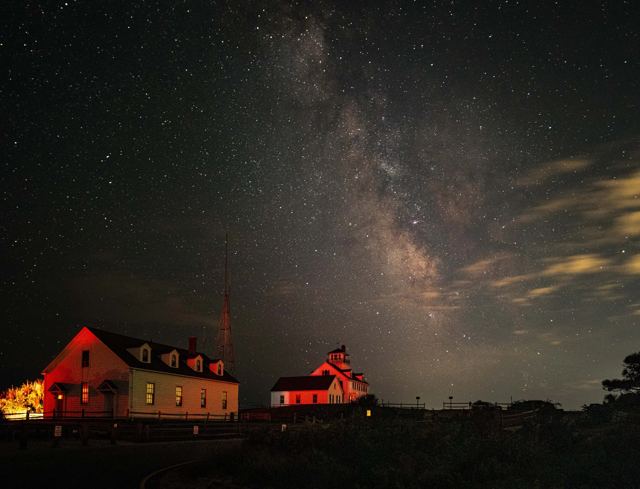 Night sky filled with stars and the Milky Way galaxy over buildings with illuminated windows, a fence, and a cloudy sky, in a rural setting.