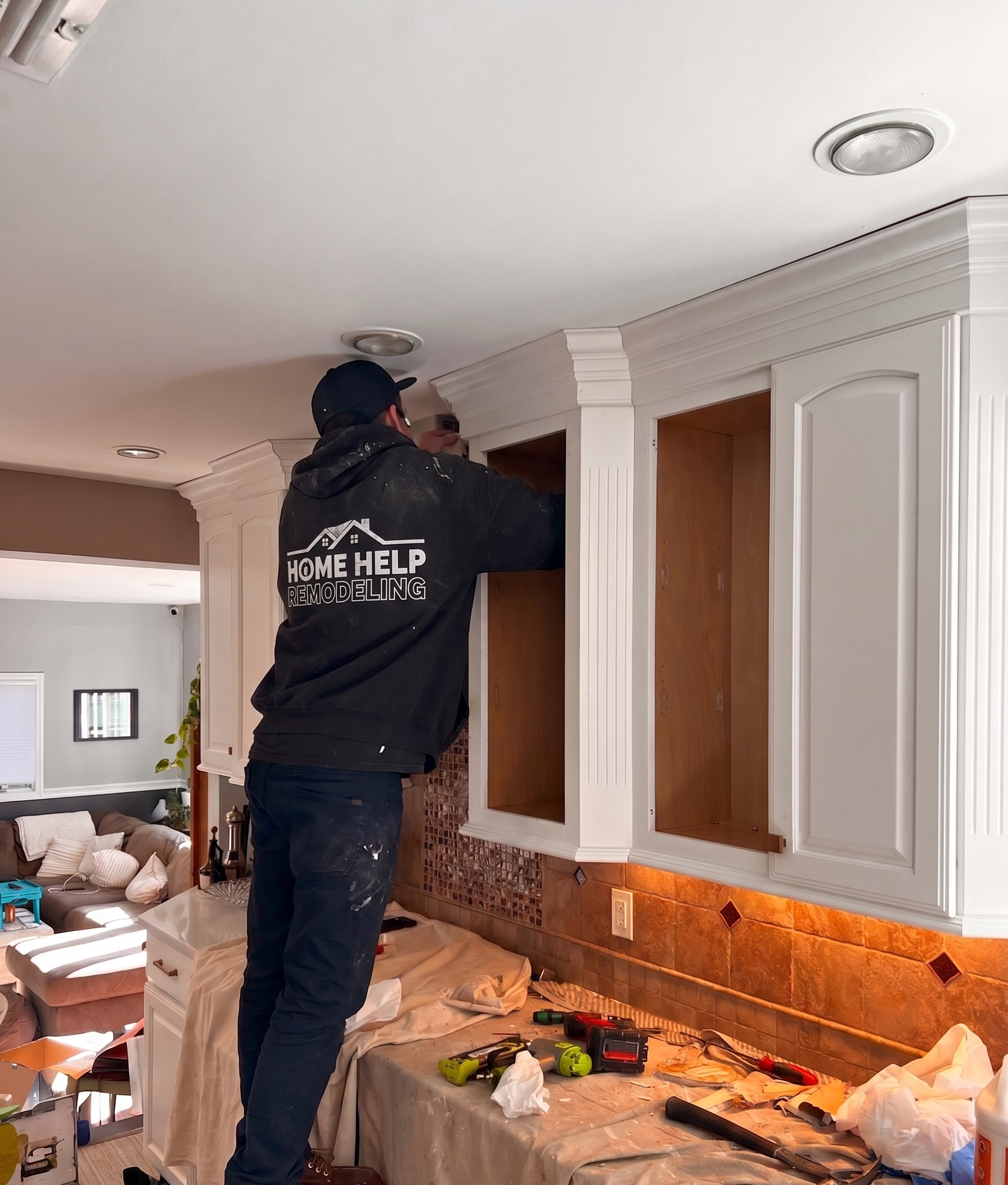 A person wearing a black hoodie and cap labeled 'Home Help Remodeling' works on installing or repairing a kitchen cabinet in a home. The workspace is cluttered with tools like drills, screwdrivers, and other equipment, with some parts of the cabinet and a plastic sheet covering the counter. The room has a cozy living area with a brown sofa, pillows, and a small table in the background.