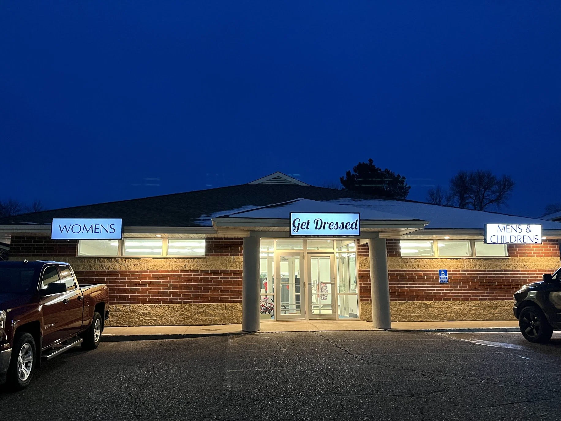 Night view of a small shopping center with three lit signs reading WOMENS, Get Dressed, and MENS & CHILDRENS. A brick building with glass entrance doors, parked cars, and a small parking lot.